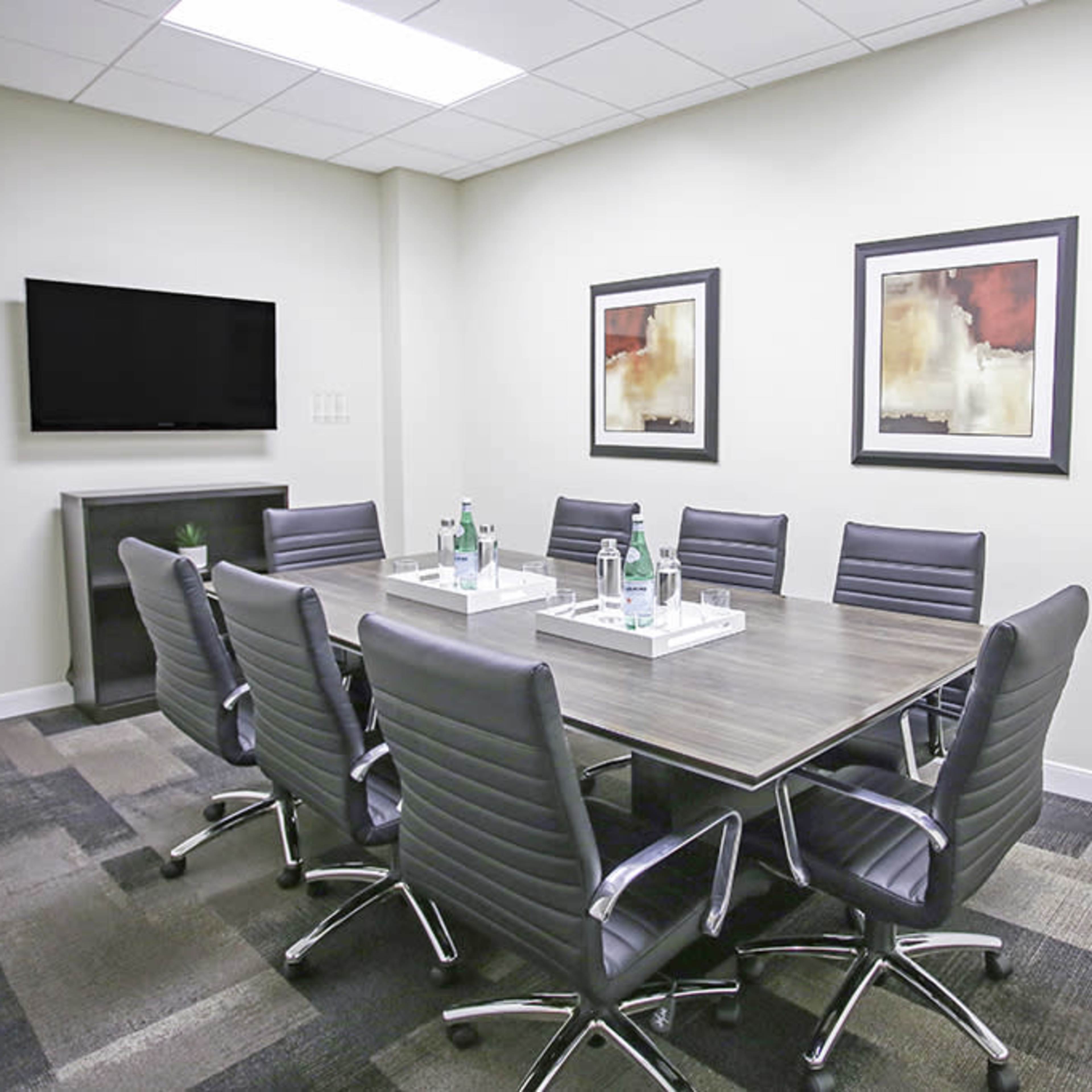 A modern conference room features a large table surrounded by eight ergonomic chairs, with two framed artworks and a television on the wall.