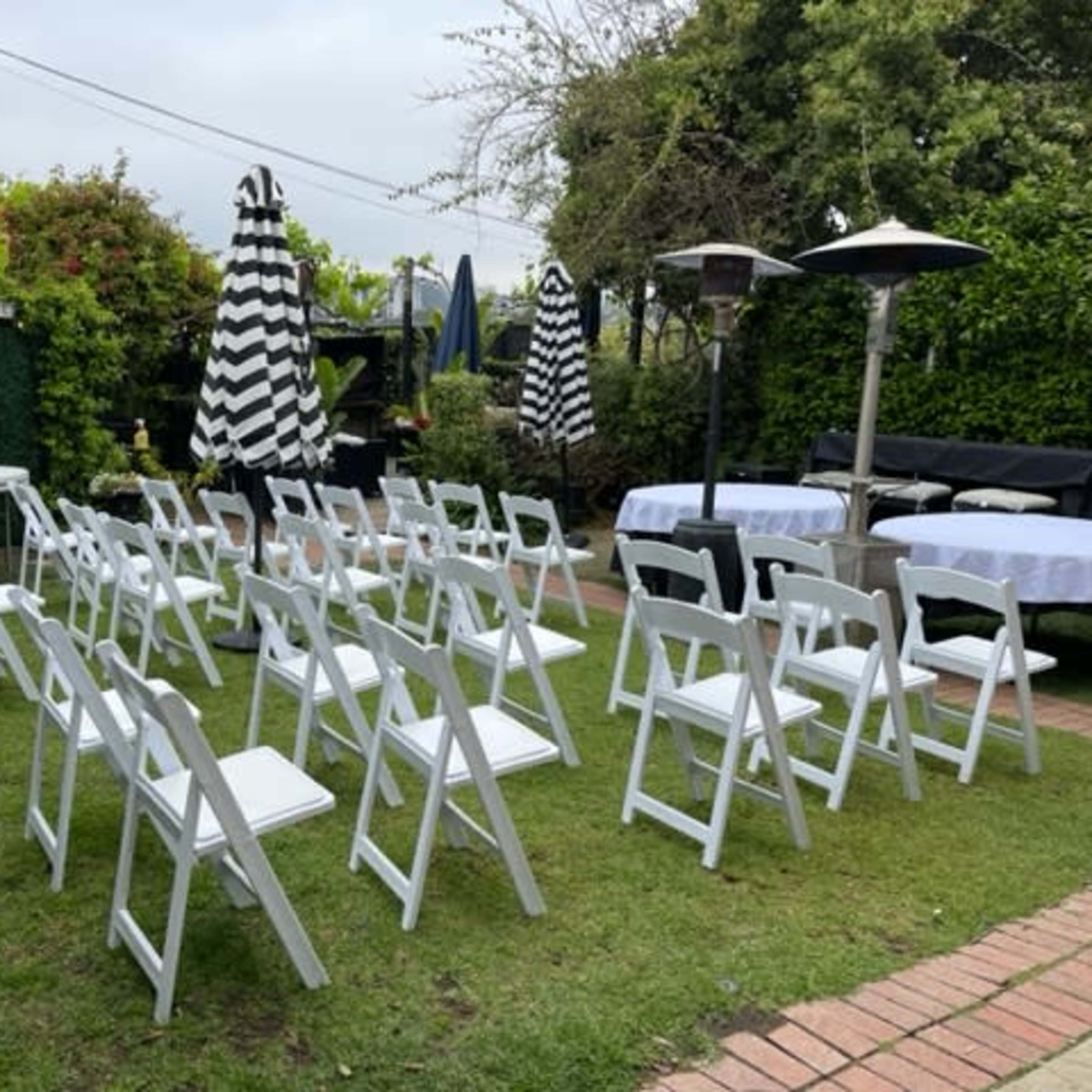 The scene shows a garden seating arrangement with white folding chairs facing a table under striped umbrellas, surrounded by greenery.