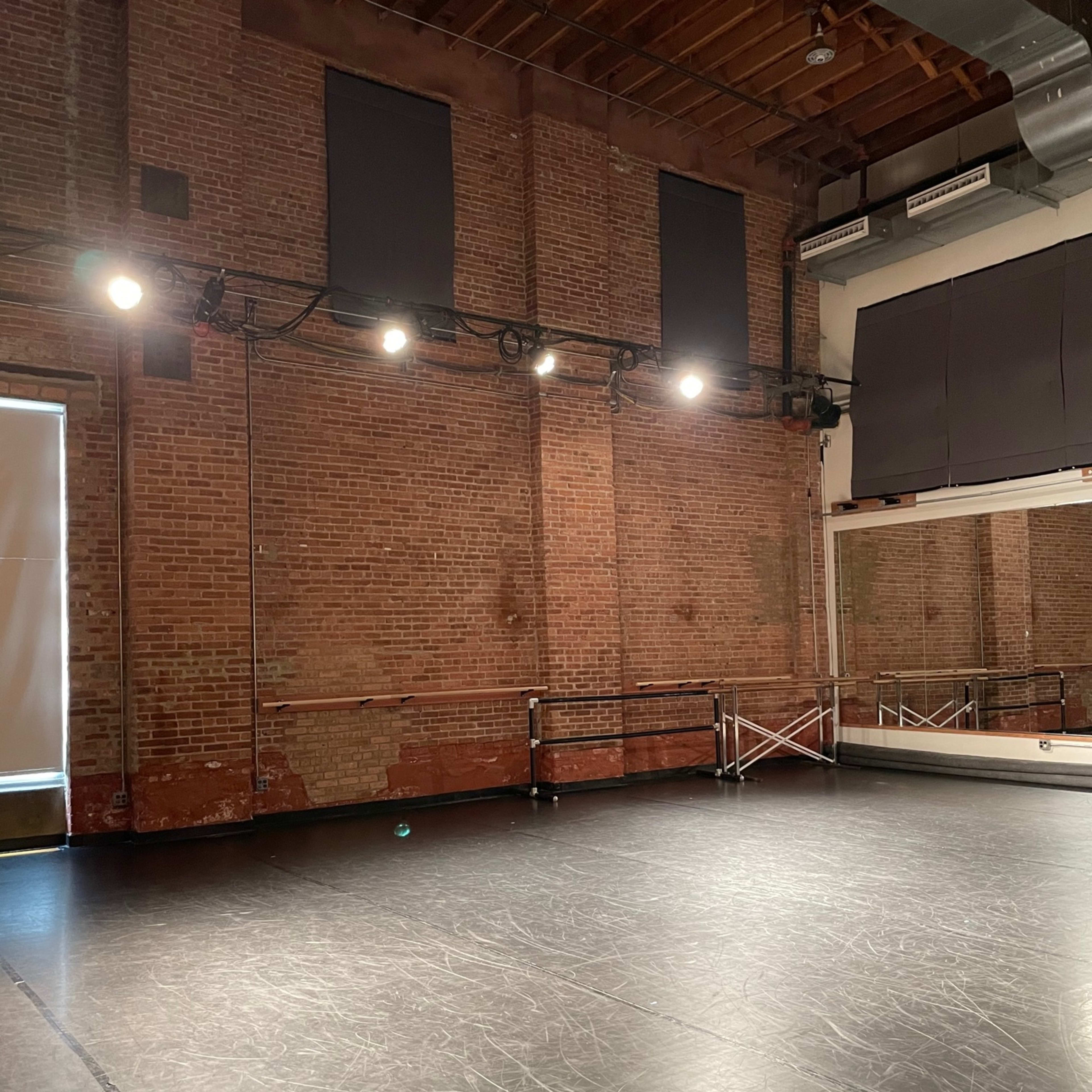 A spacious dance studio with exposed brick walls, overhead lights, and a mirrored wall.
