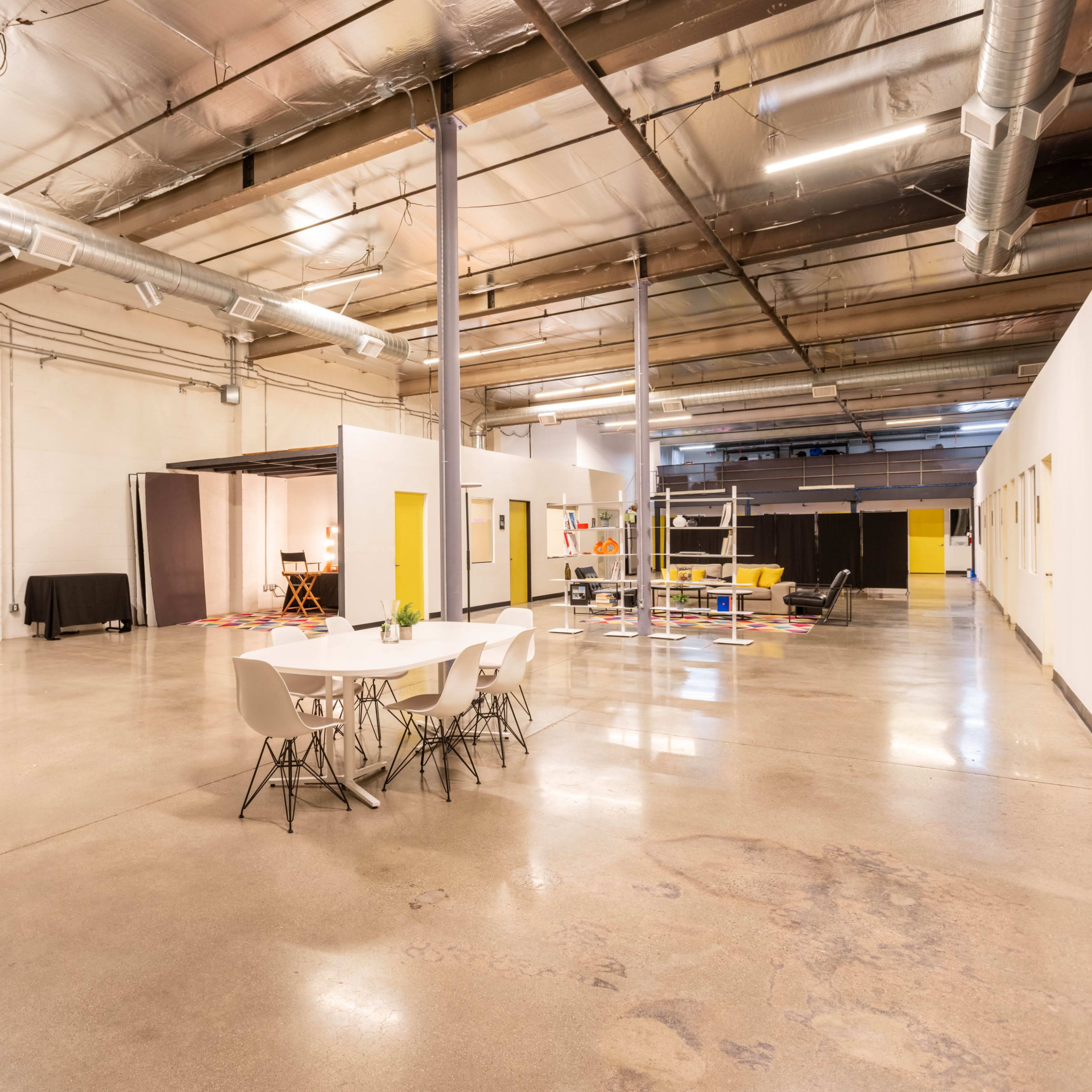 A spacious, well-lit industrial interior featuring a dining table with chairs, empty areas, and various rooms with yellow doors.