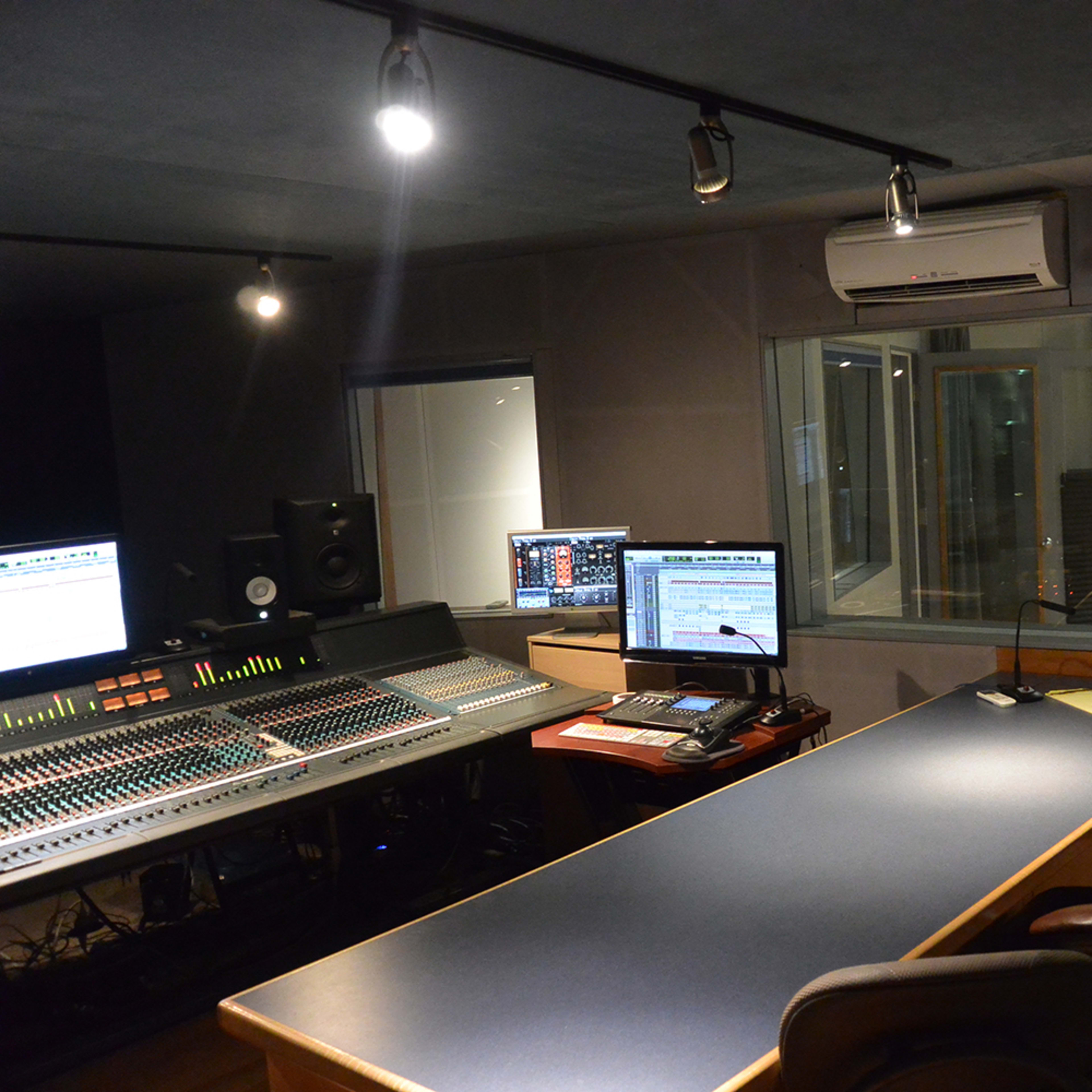 A music recording studio with an audio mixing console, computer monitors, and a large desk, featuring wooden floors and a separate room visible through glass windows.