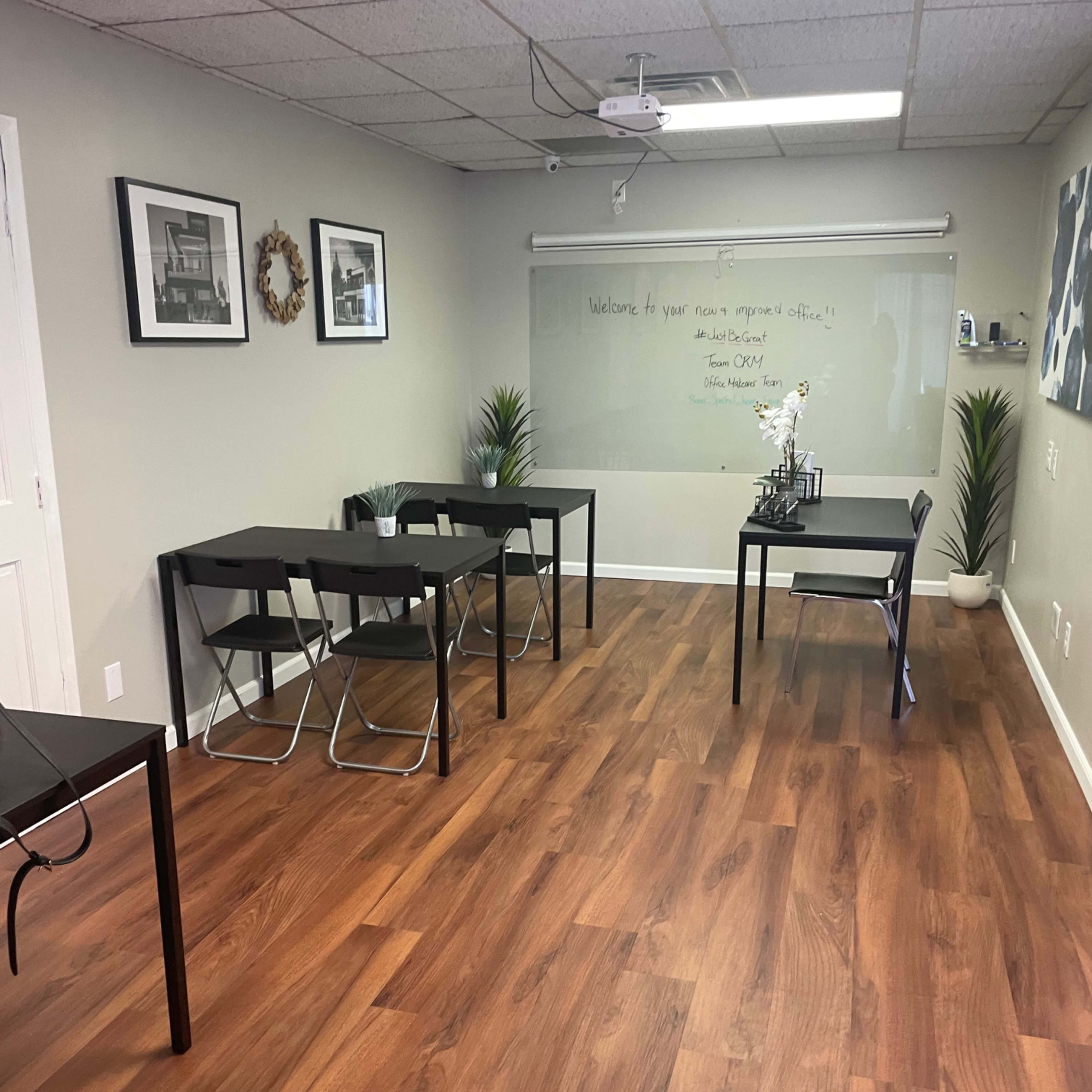 A room with wooden flooring, featuring several black tables and chairs, a whiteboard with a welcome message, and wall decorations.