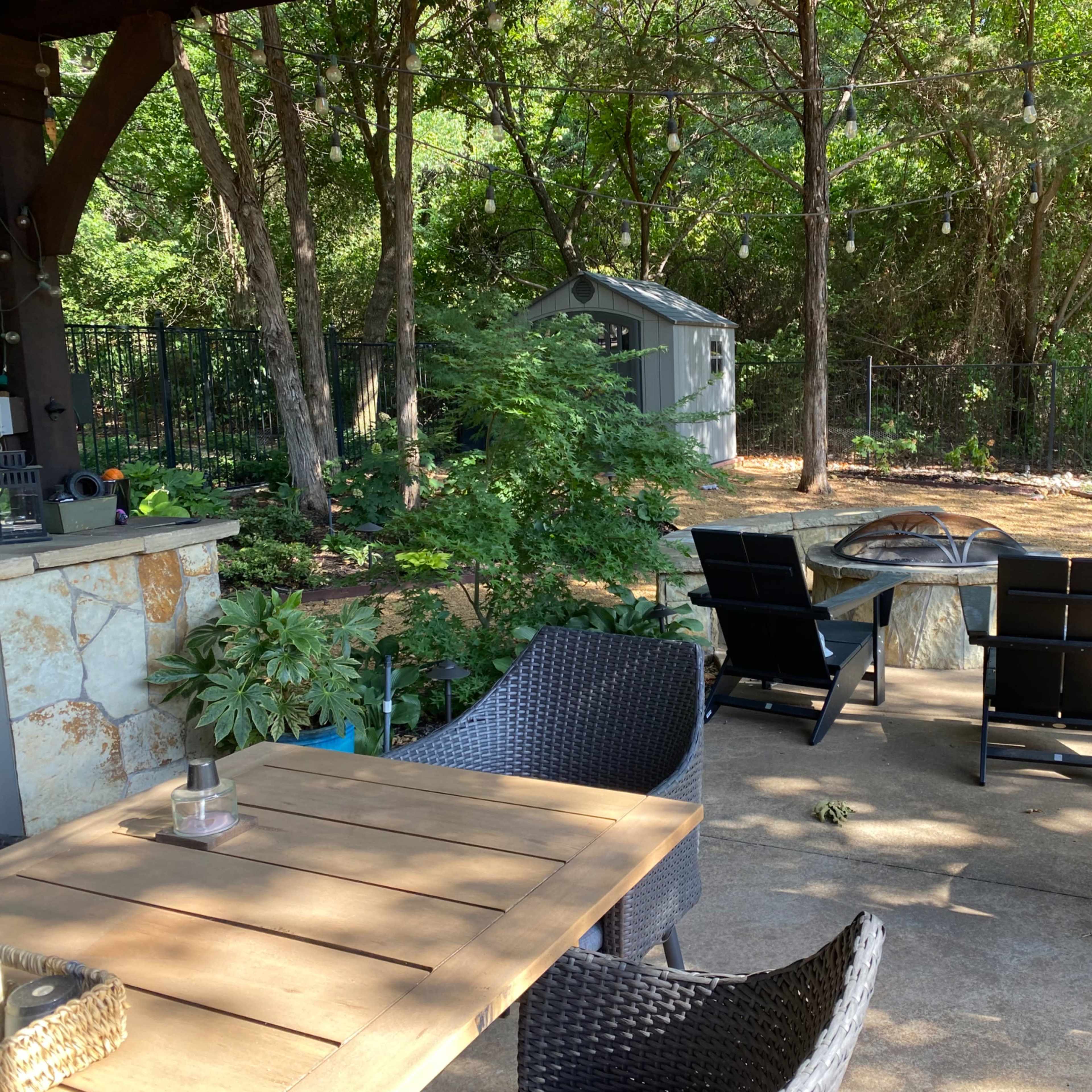 A patio area with a stone table and chairs overlooks a landscaped yard featuring trees and a shed.