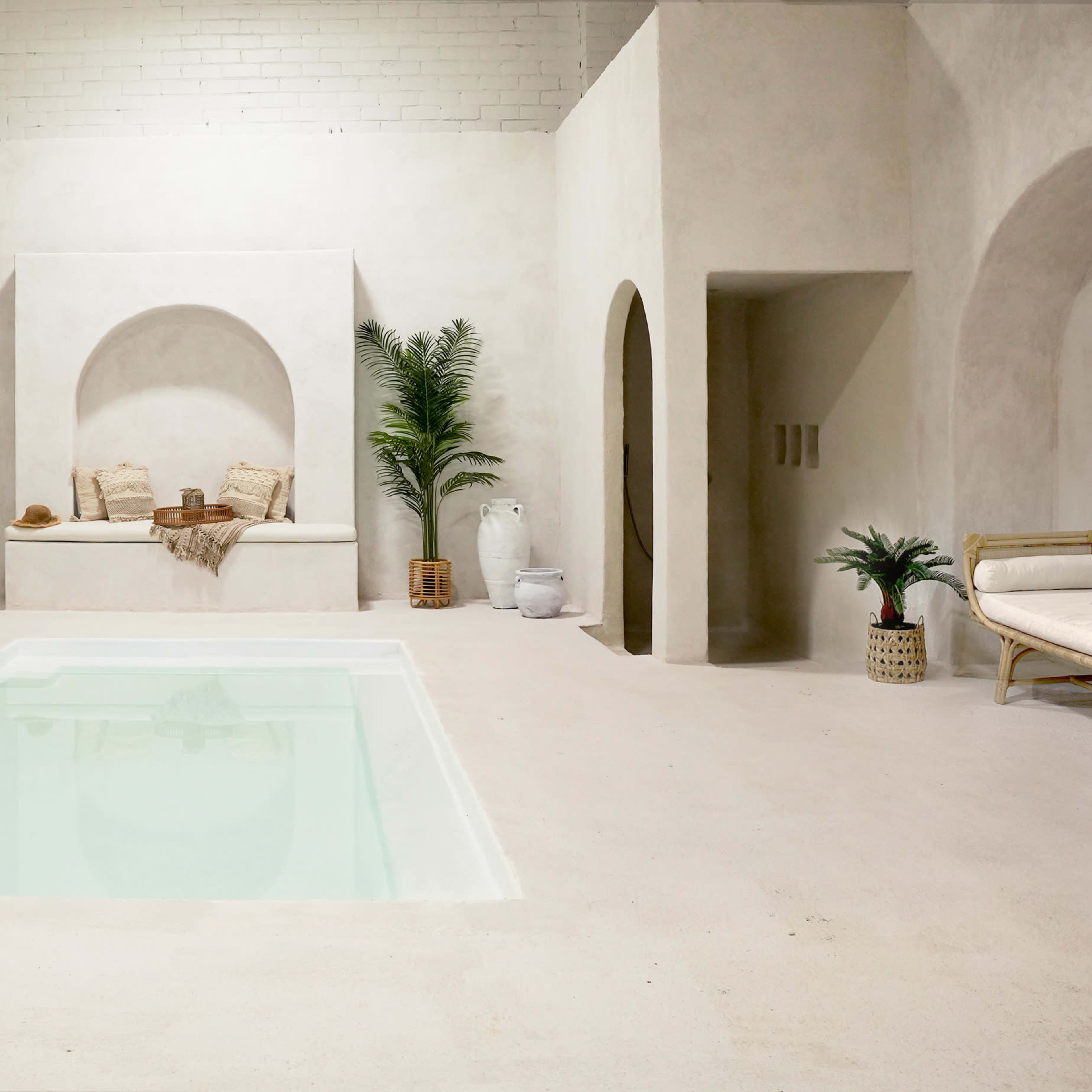 A minimalist interior space featuring a small pool, white walls, and several archways, with potted plants and seating arrangements.