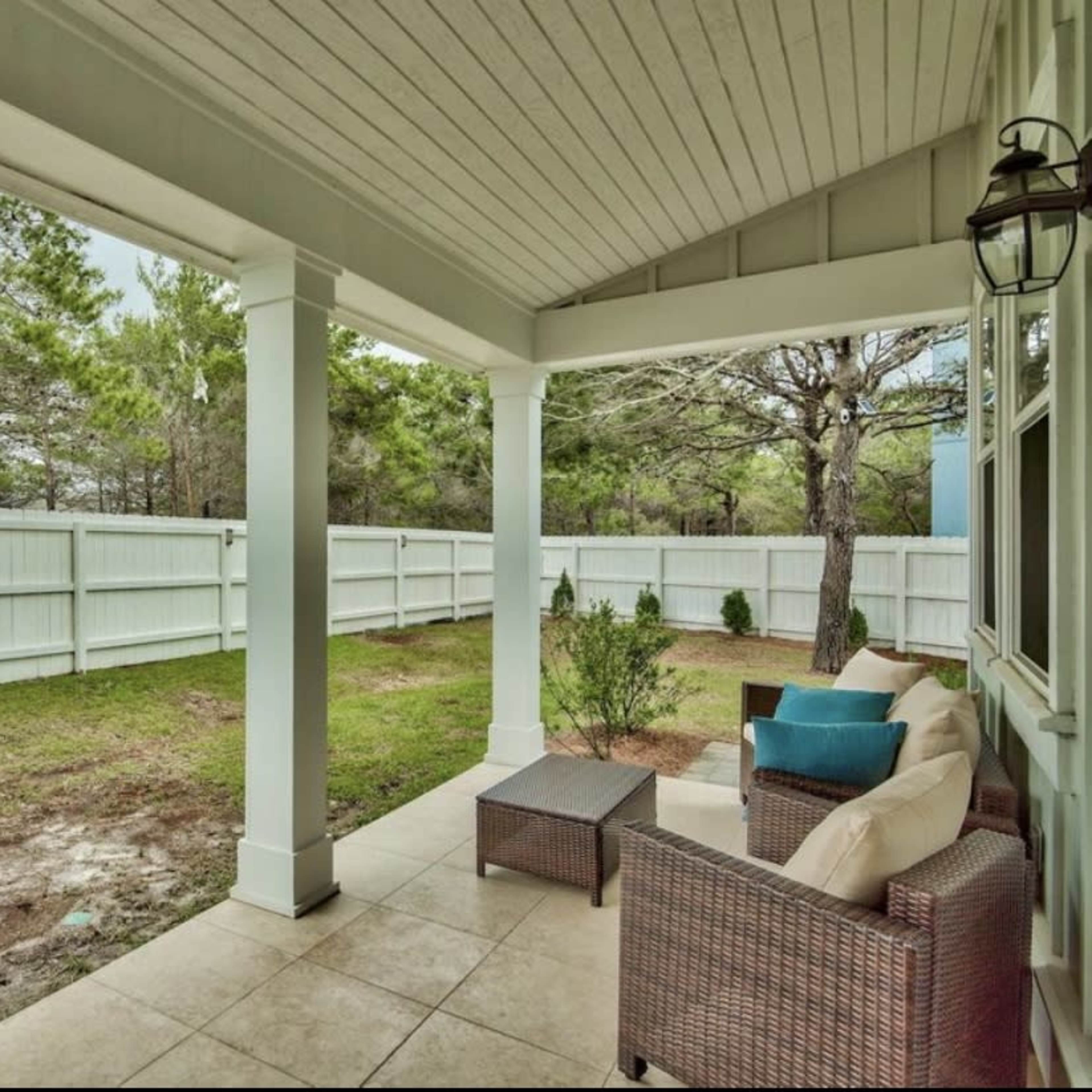 A covered patio with a seating area overlooks a grassy yard enclosed by a white fence.