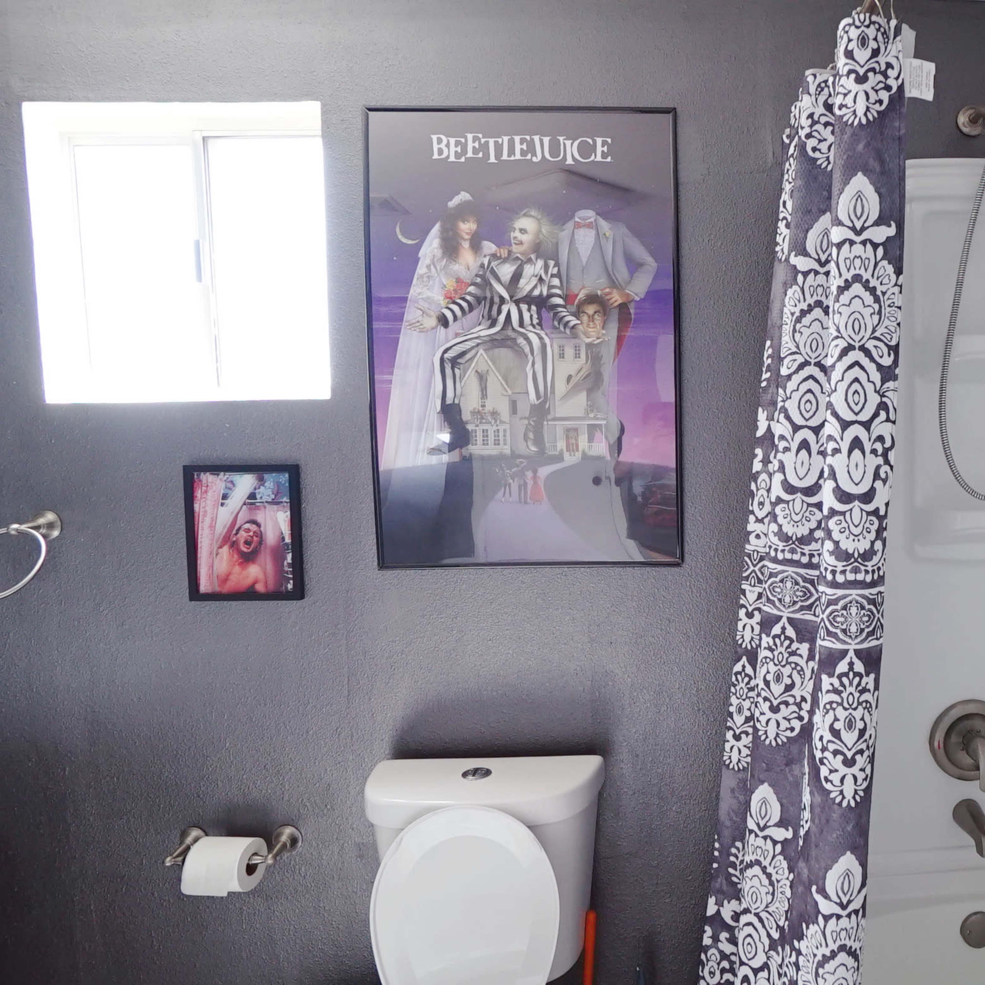 The bathroom features a shower with a patterned curtain, a toilet, a framed poster of "Beetlejuice" on the wall, and a small window allowing natural light.