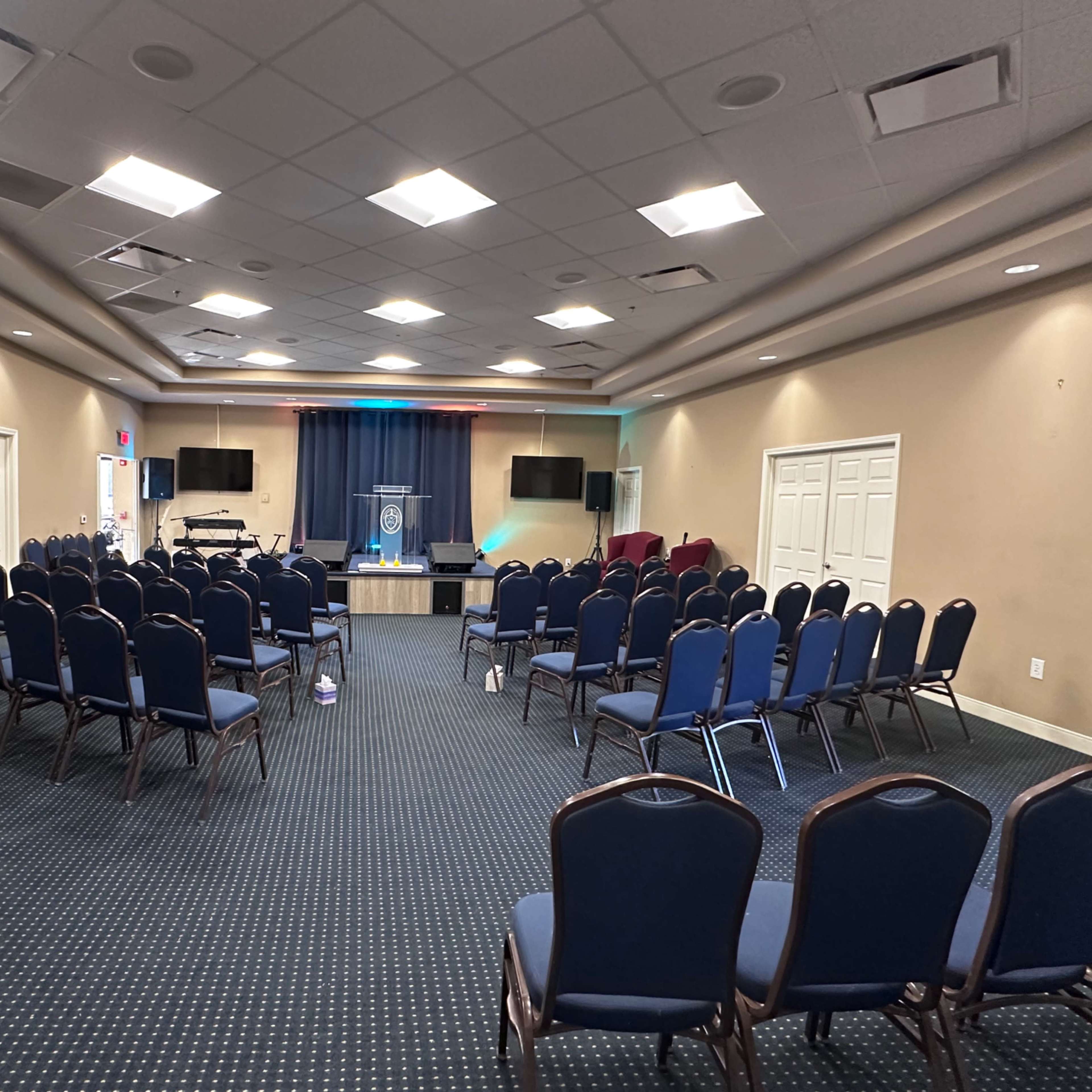 A large meeting room is set with rows of blue chairs facing a stage, with a podium at the front and a backdrop featuring a logo.