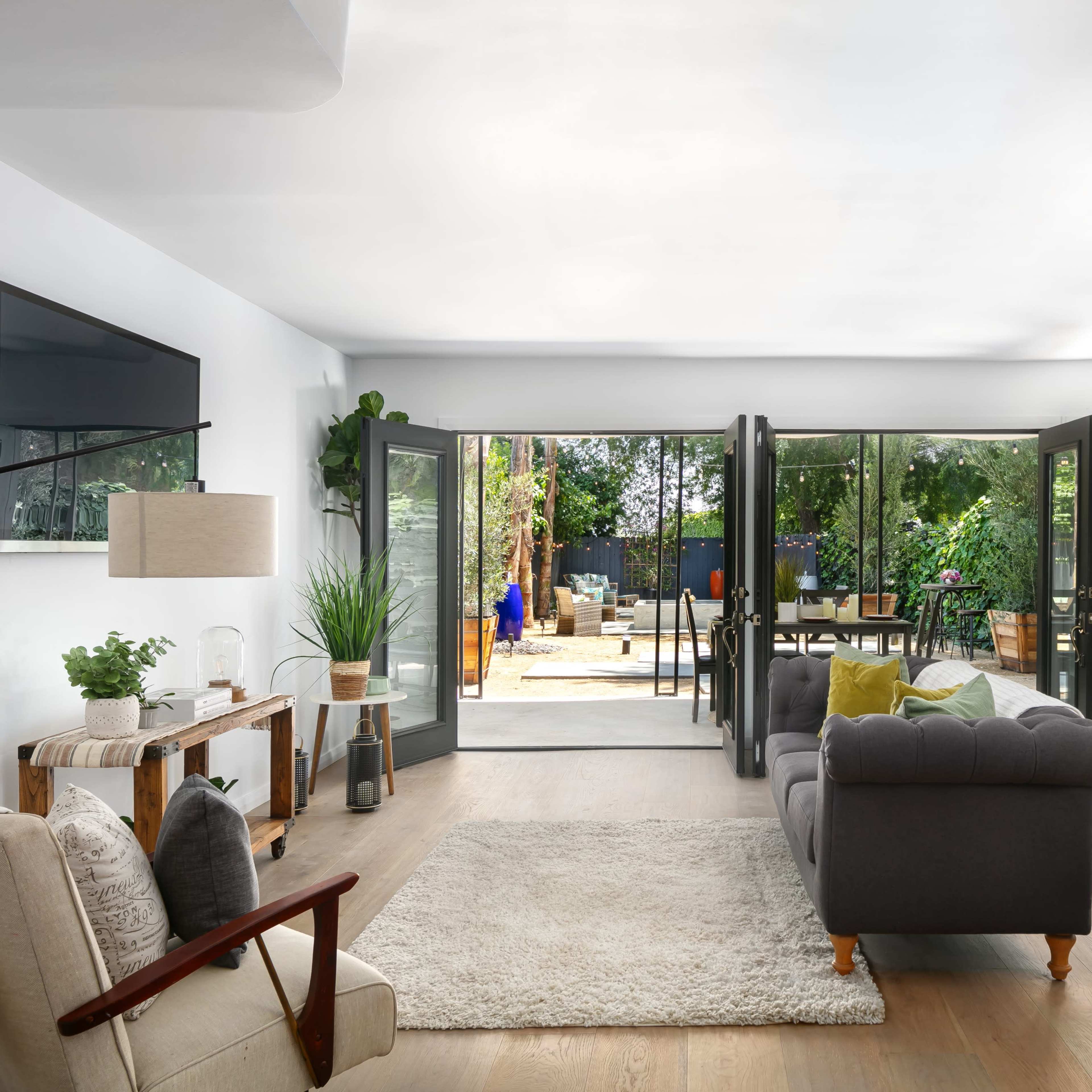 A modern living room opens up to a garden through large glass doors, featuring a sofa, an armchair, and a dining area visible in the background.