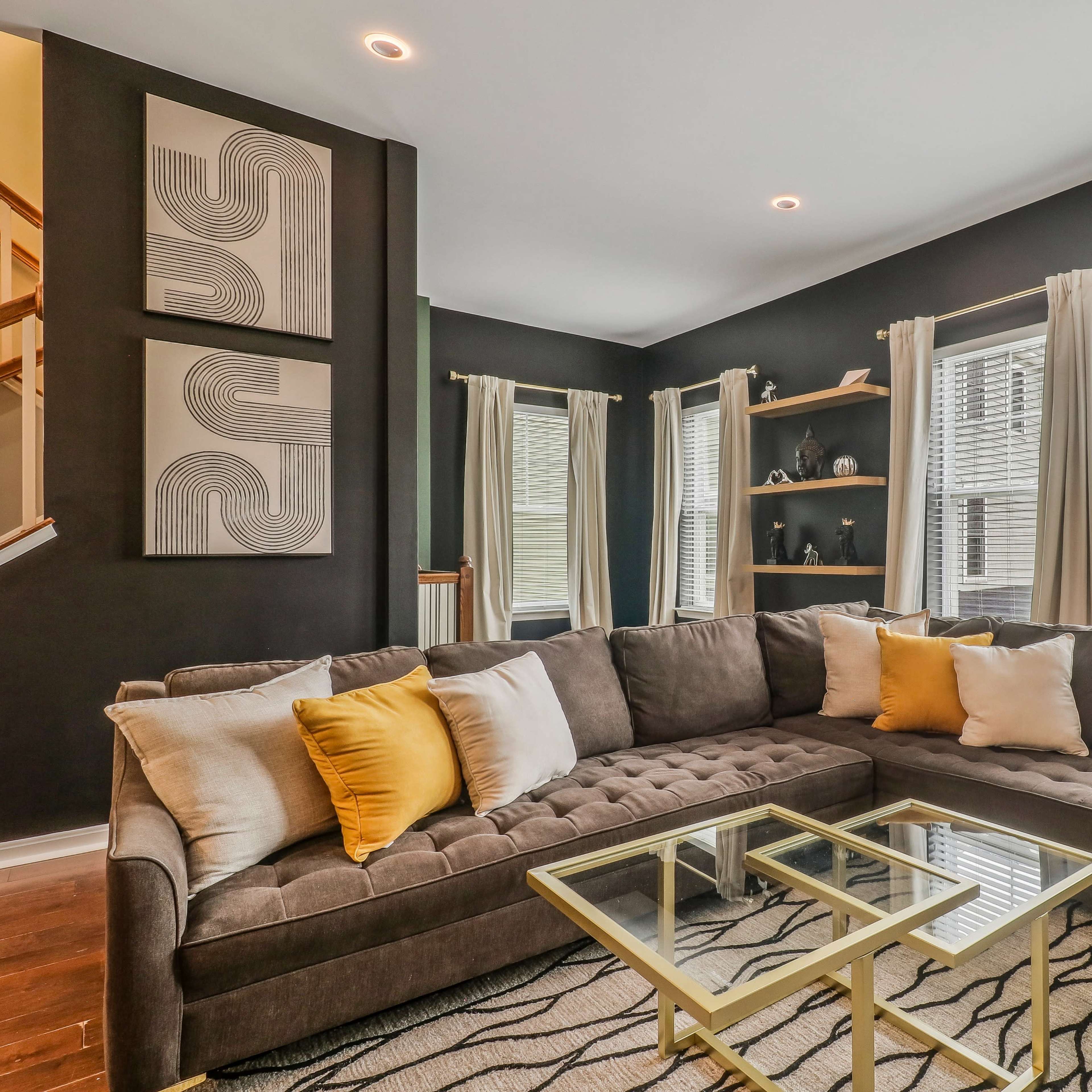 A modern living room with a gray sectional sofa adorned with yellow and white pillows, situated near large windows and decorated with wall art and shelves.