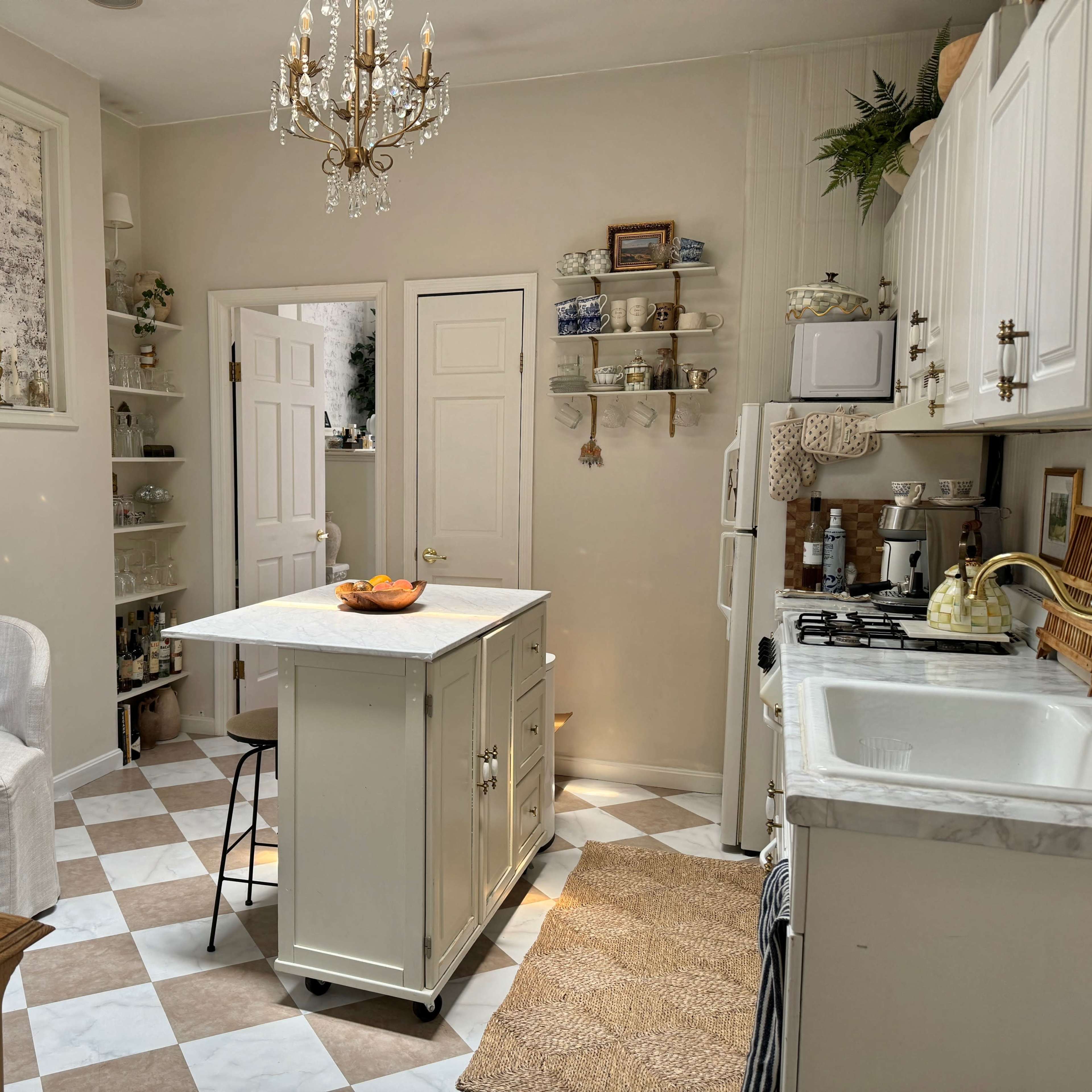 A well-organized kitchen features white cabinets, a central island with a bowl of fruit, and checkered flooring with shelves displaying decorative items.