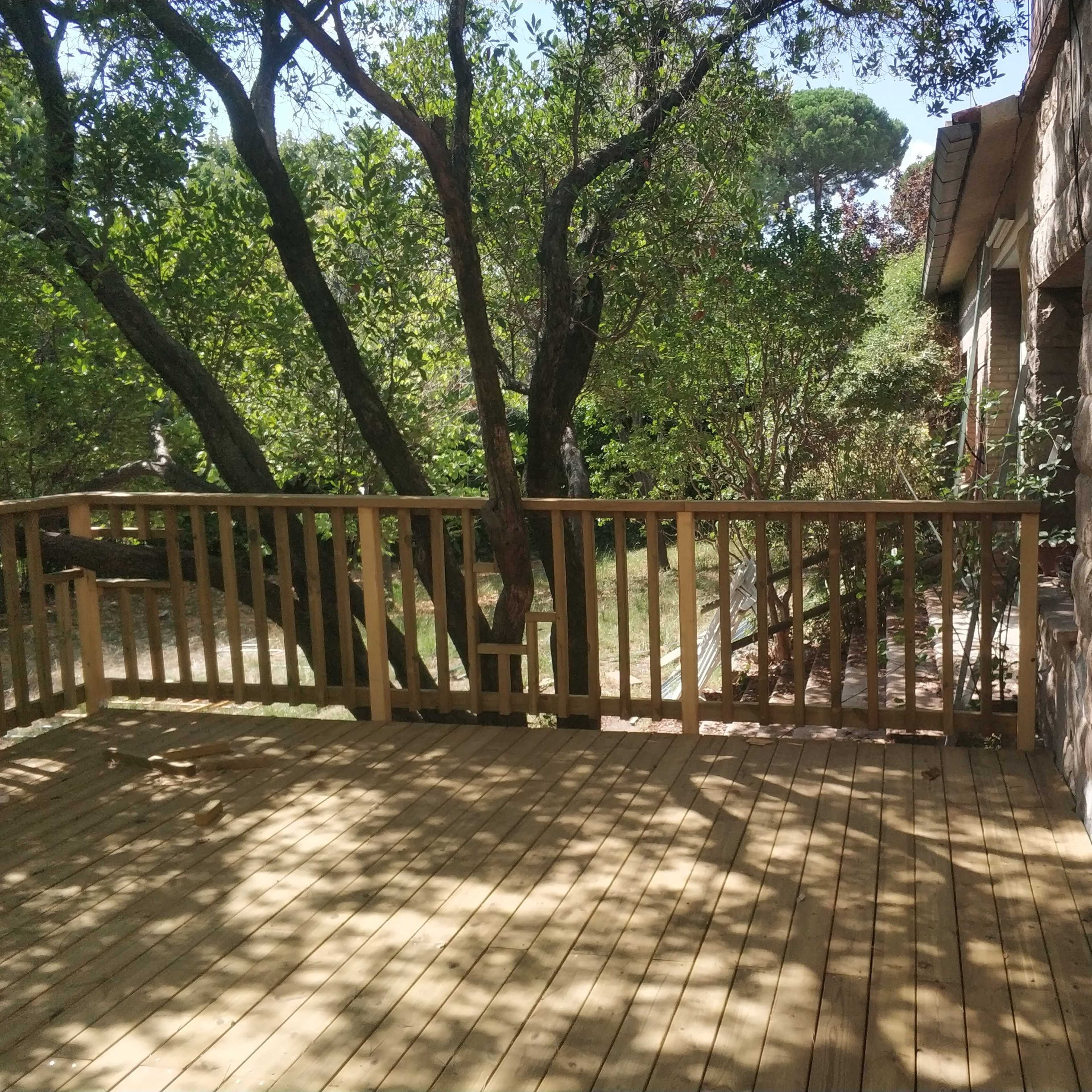 A wooden deck surrounded by trees and greenery leads to a path beside a house.