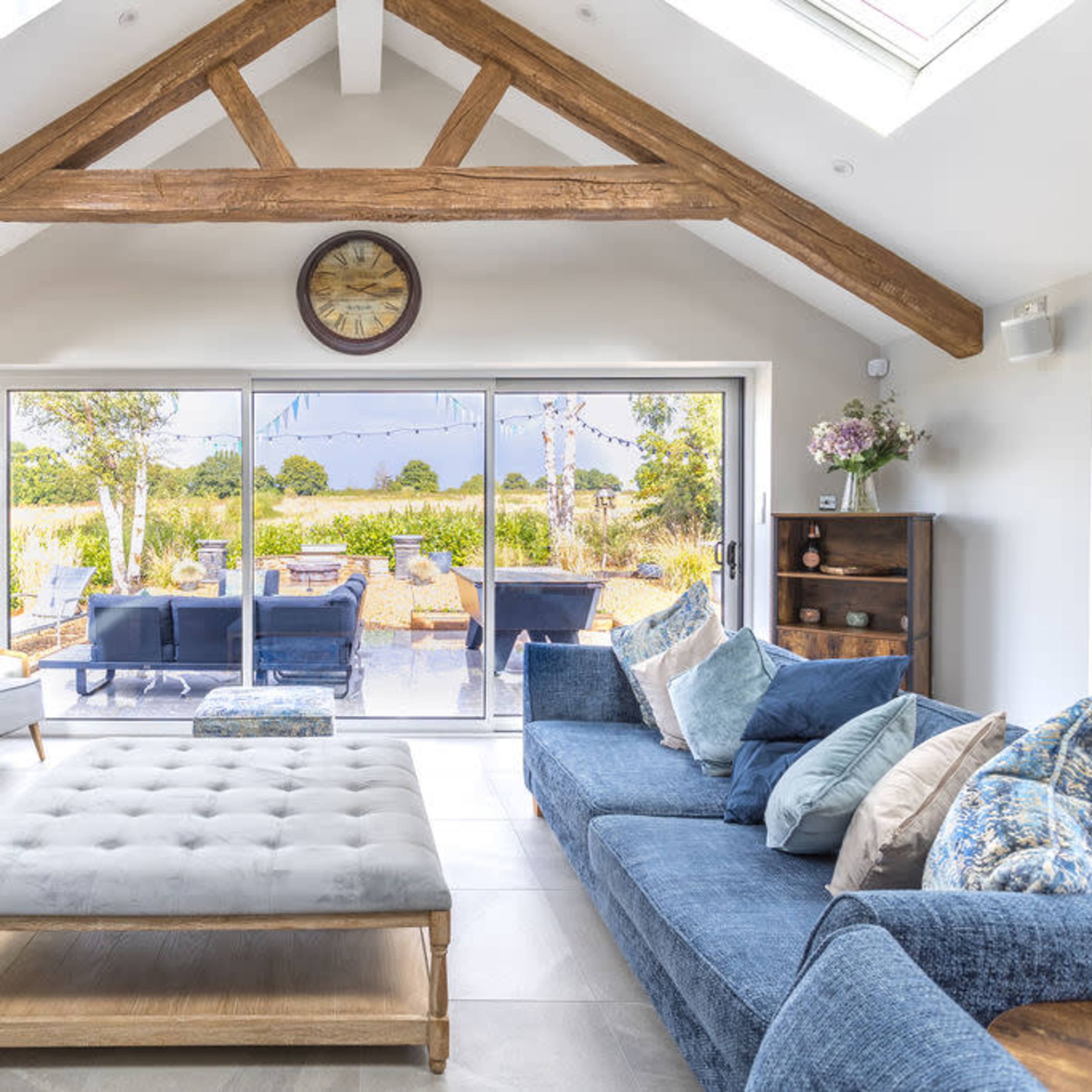 A modern living room features a large sectional sofa, a coffee table, and a flat-screen television, with sliding glass doors opening to a view of an outdoor area with seating and greenery.