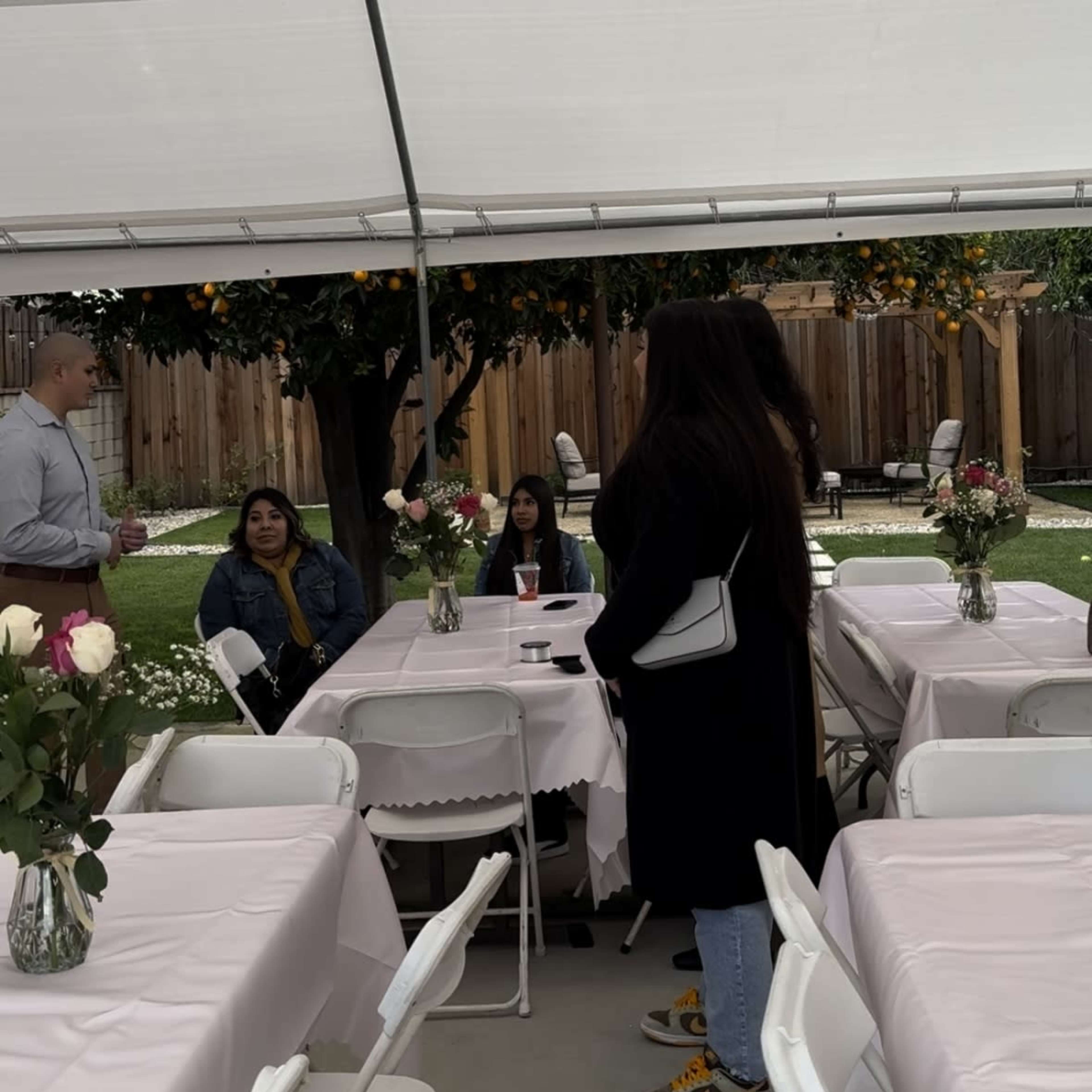A group of people sits at tables under a canopy, with flowers placed in vases on the tables and an orange tree visible in the background.