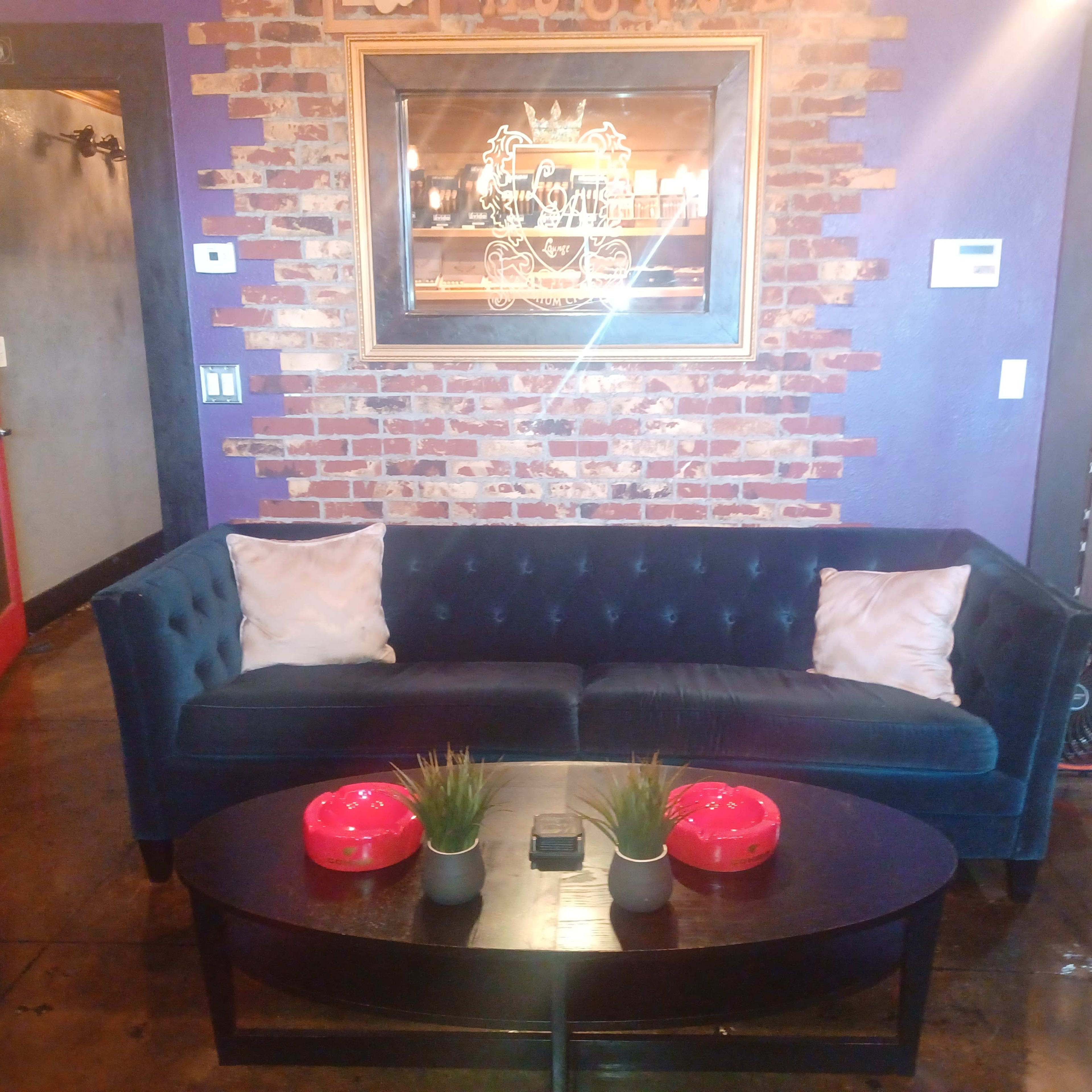 A dark blue velvet sofa with two decorative pillows facing a round black coffee table, set against a brick wall with framed artwork above.