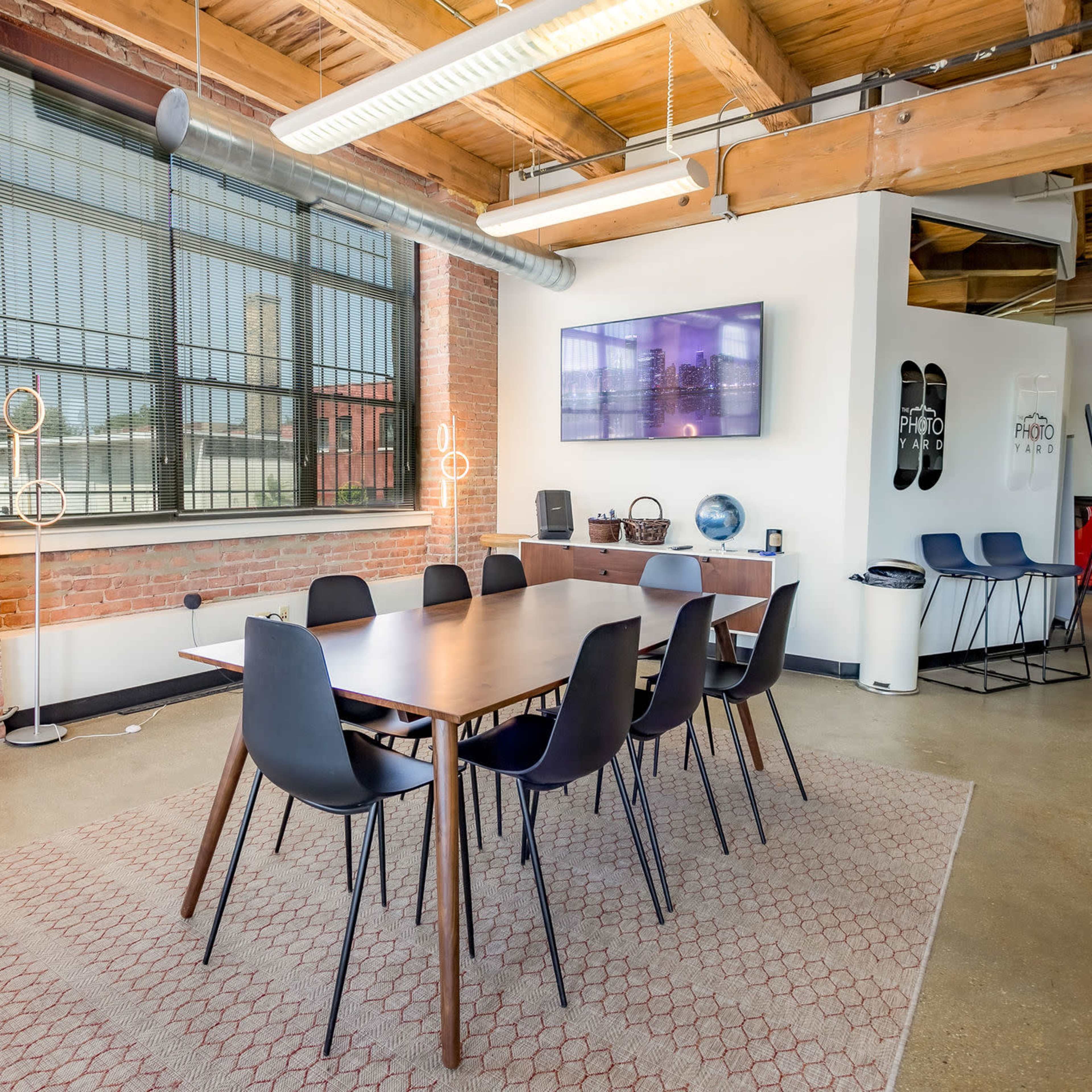 A conference room features a long wooden table surrounded by black chairs, with exposed brick walls and large windows.