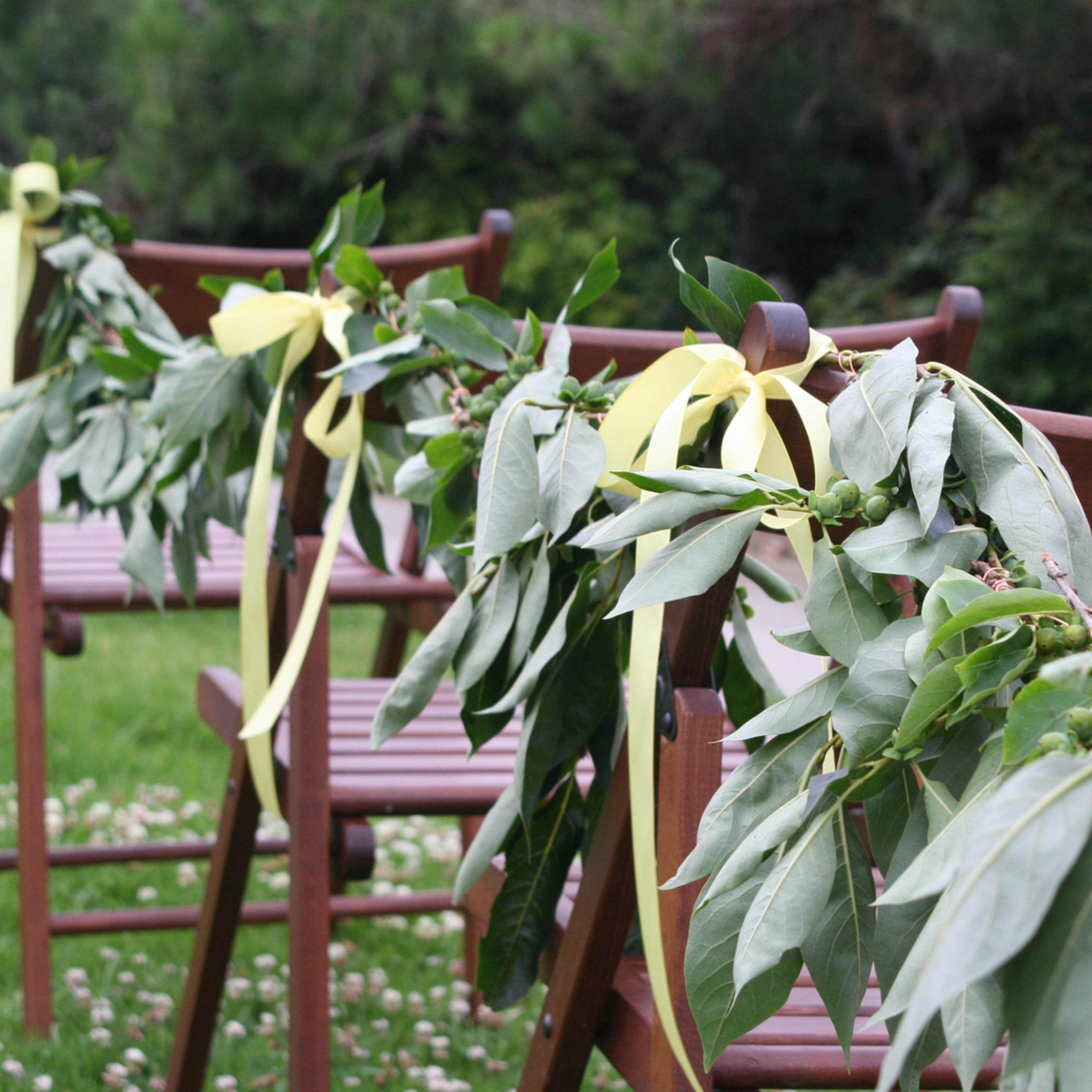 Wooden chairs are decorated with leafy garlands and yellow ribbons in a grassy outdoor setting.