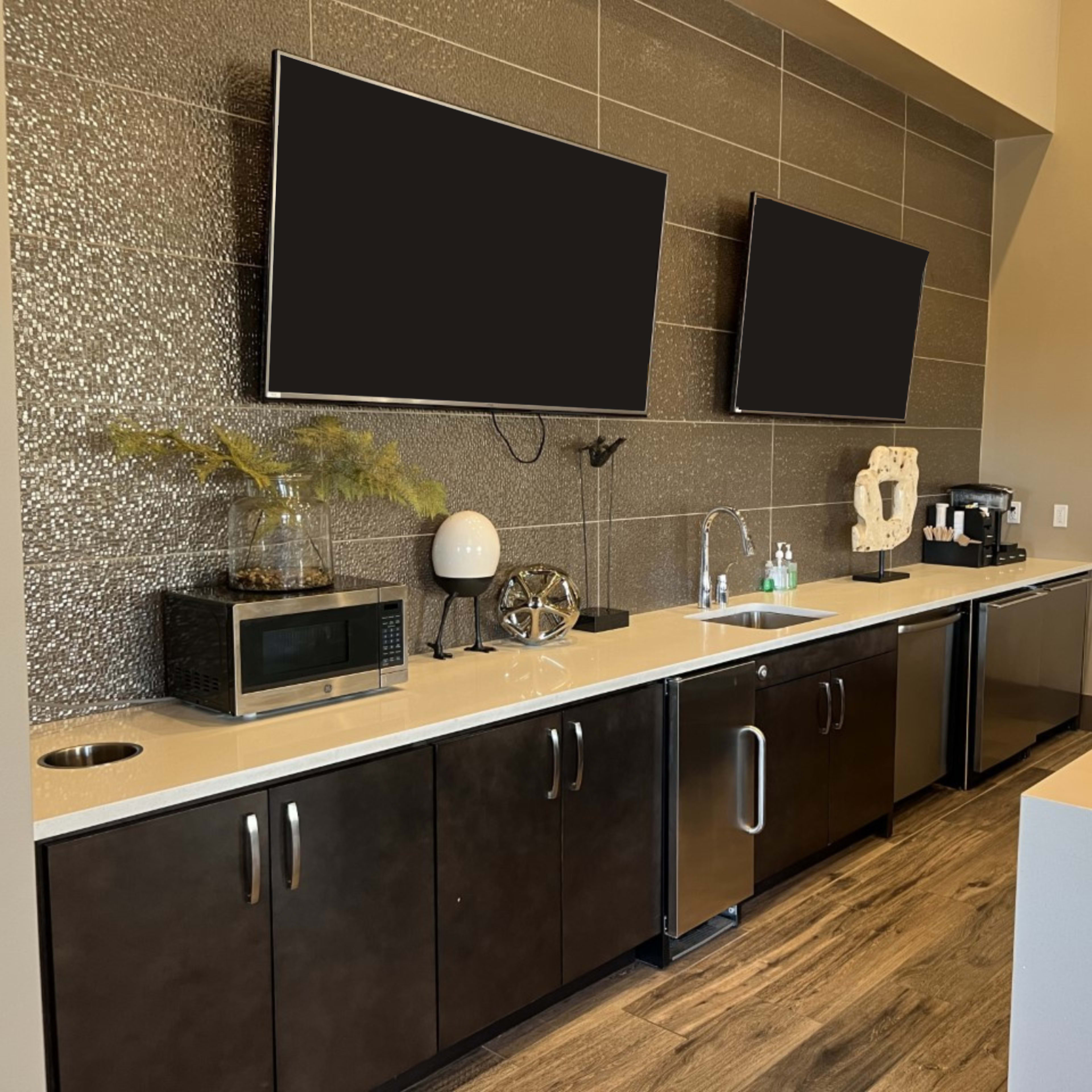 A modern kitchenette area featuring two mounted flat-screen TVs, a microwave, a coffee machine, and cabinets beneath a counter.