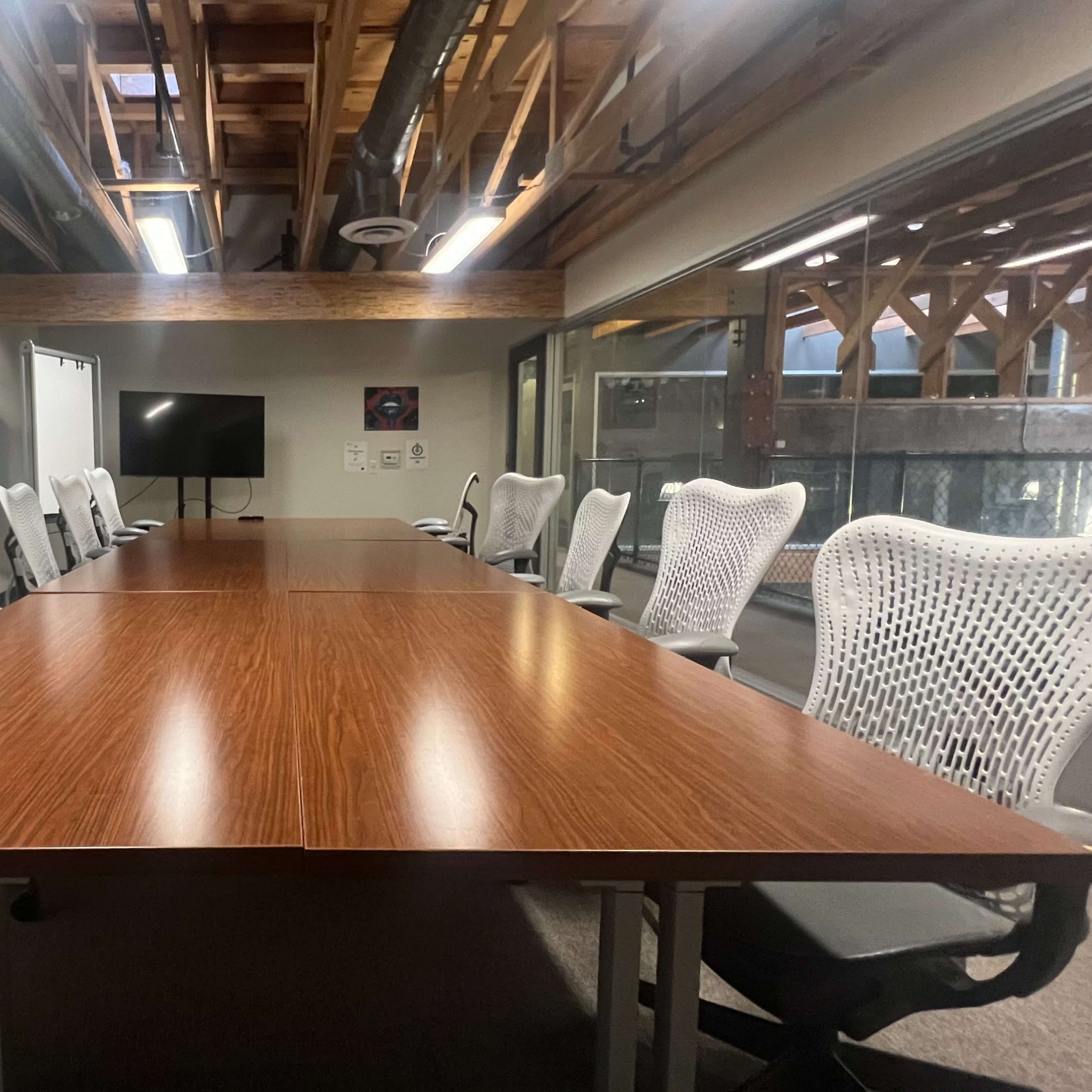 A conference room features a long wooden table surrounded by ergonomic white chairs under a ceiling with exposed beams.