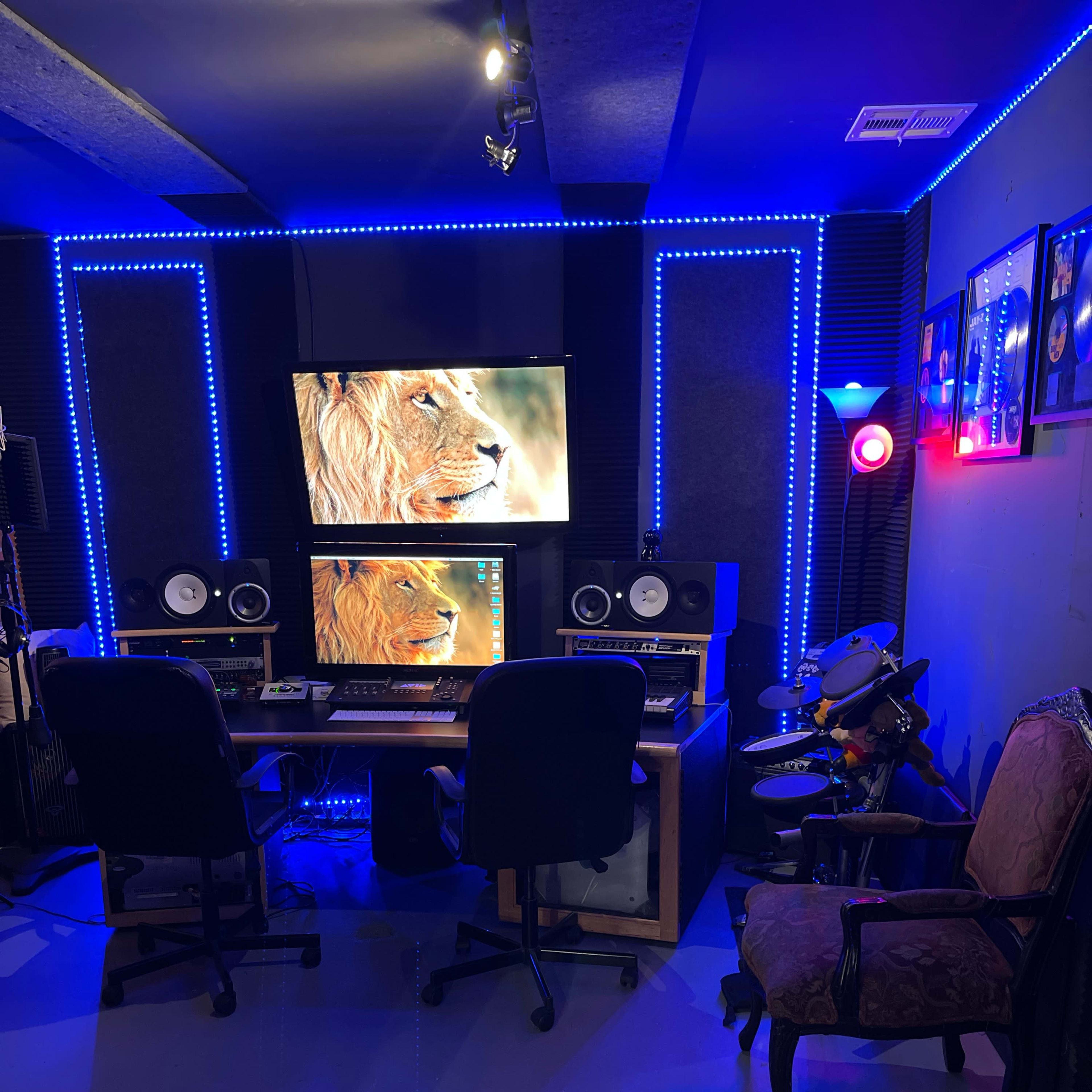 A music studio with a computer desk, two chairs, and equipment surrounded by blue LED lights and decorated walls.