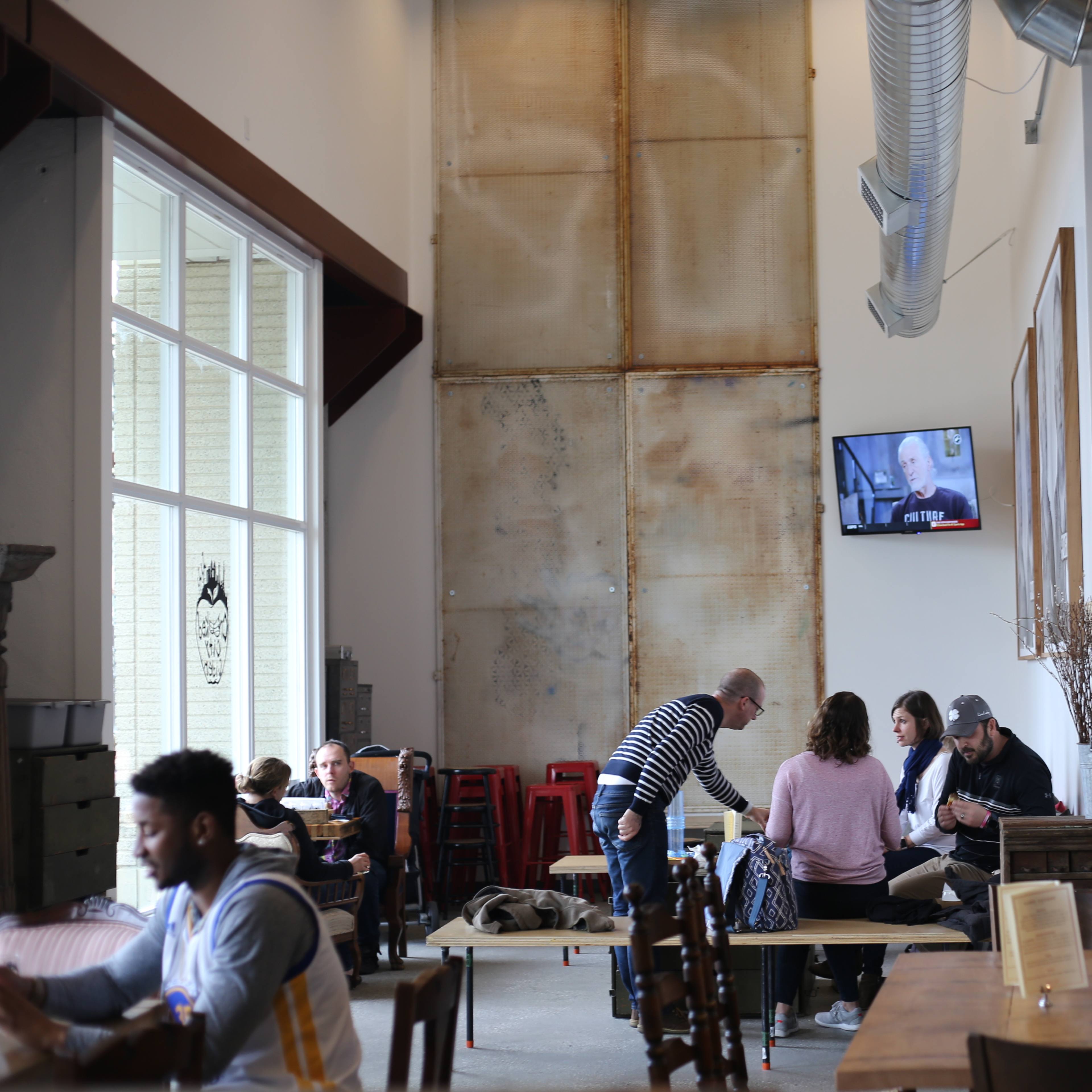 The interior of a café features a mix of tables and chairs, with patrons engaged in conversation and a television screen displaying a program on the wall.