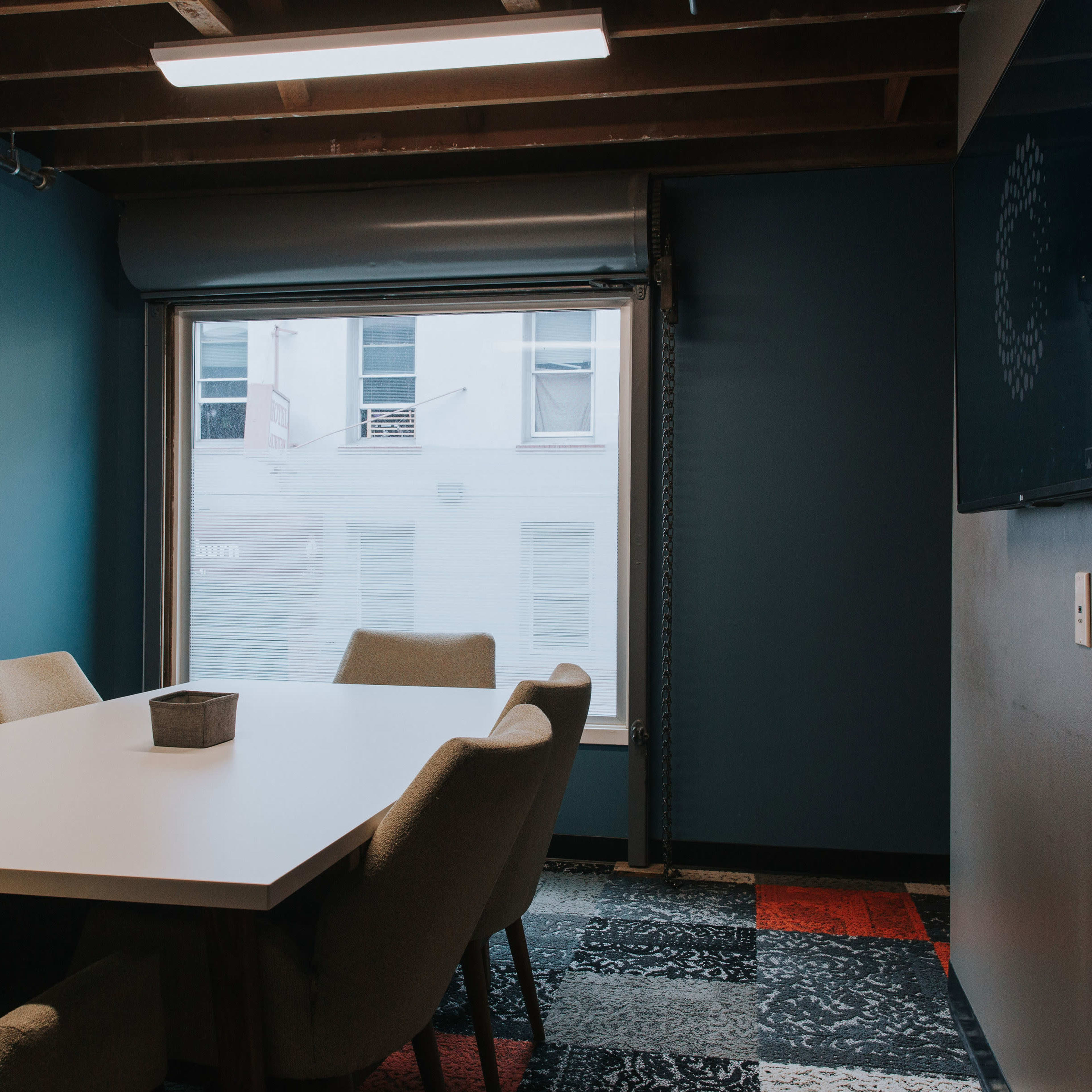 A meeting room features a rectangular white table surrounded by beige chairs, with a window showing a view of the building outside.