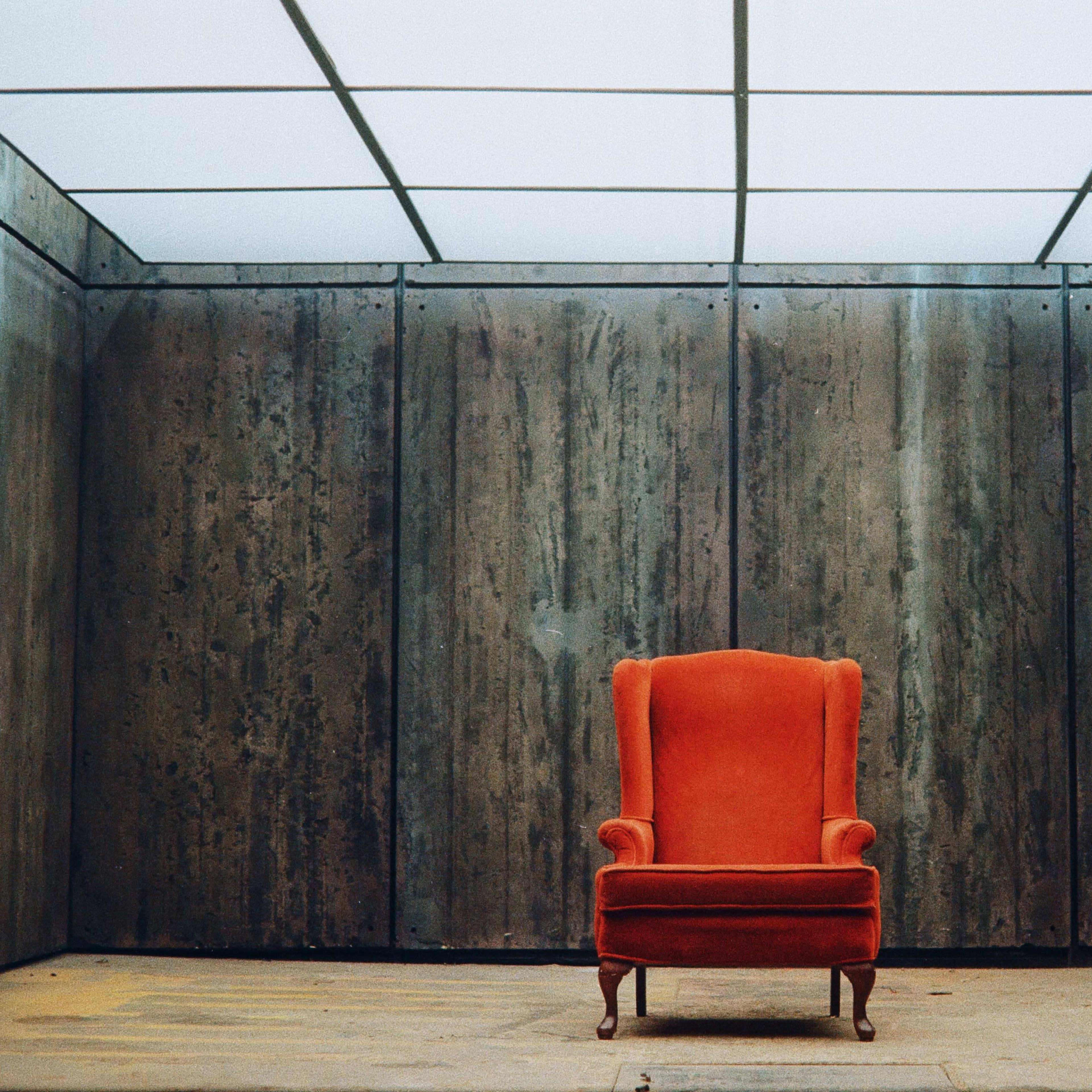 An orange armchair is positioned in the center of a minimalist, concrete-walled room with a glass ceiling.