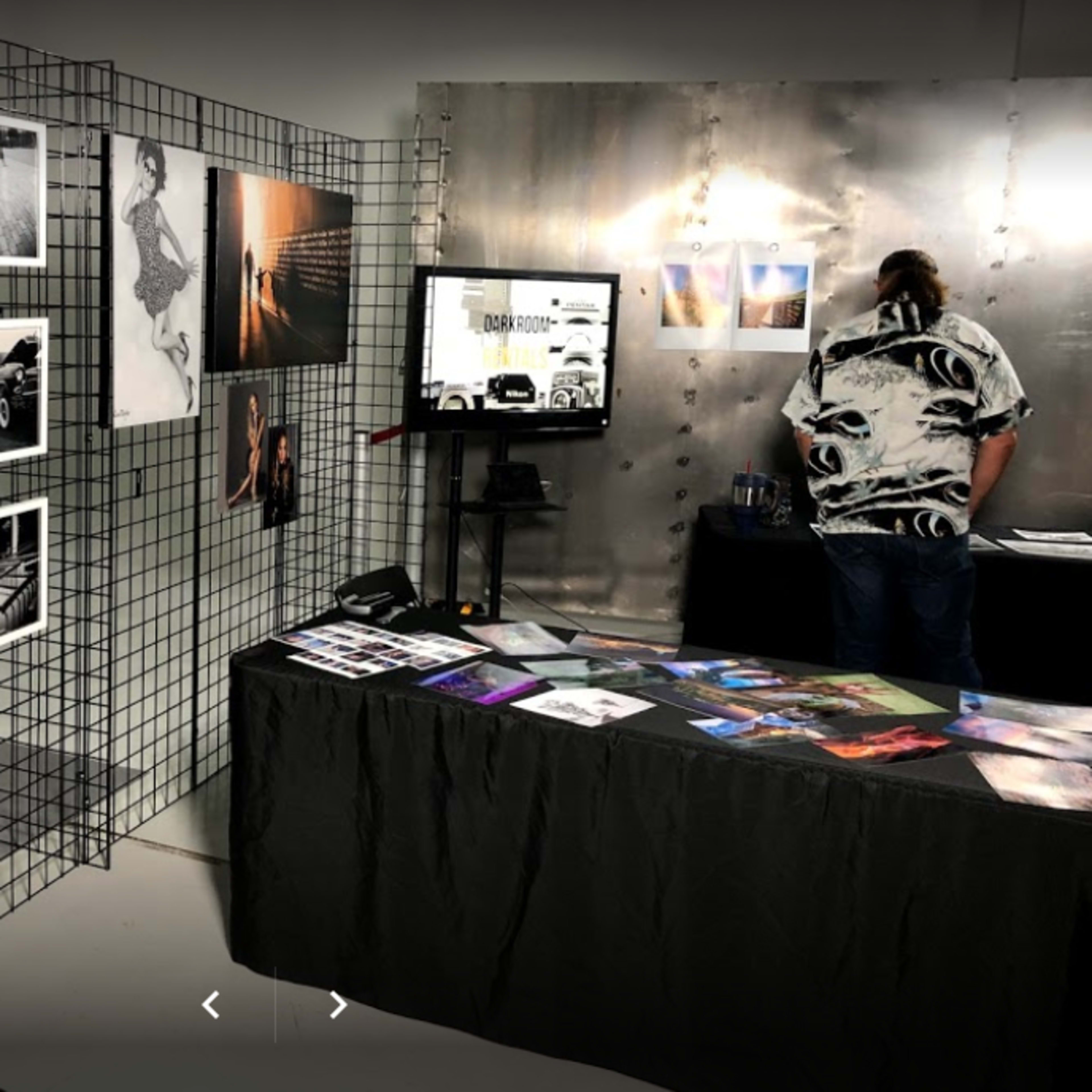 A gallery display with black-and-white photographs on a grid wall and a table covered with various printed materials, while a person stands behind the table looking at a screen.