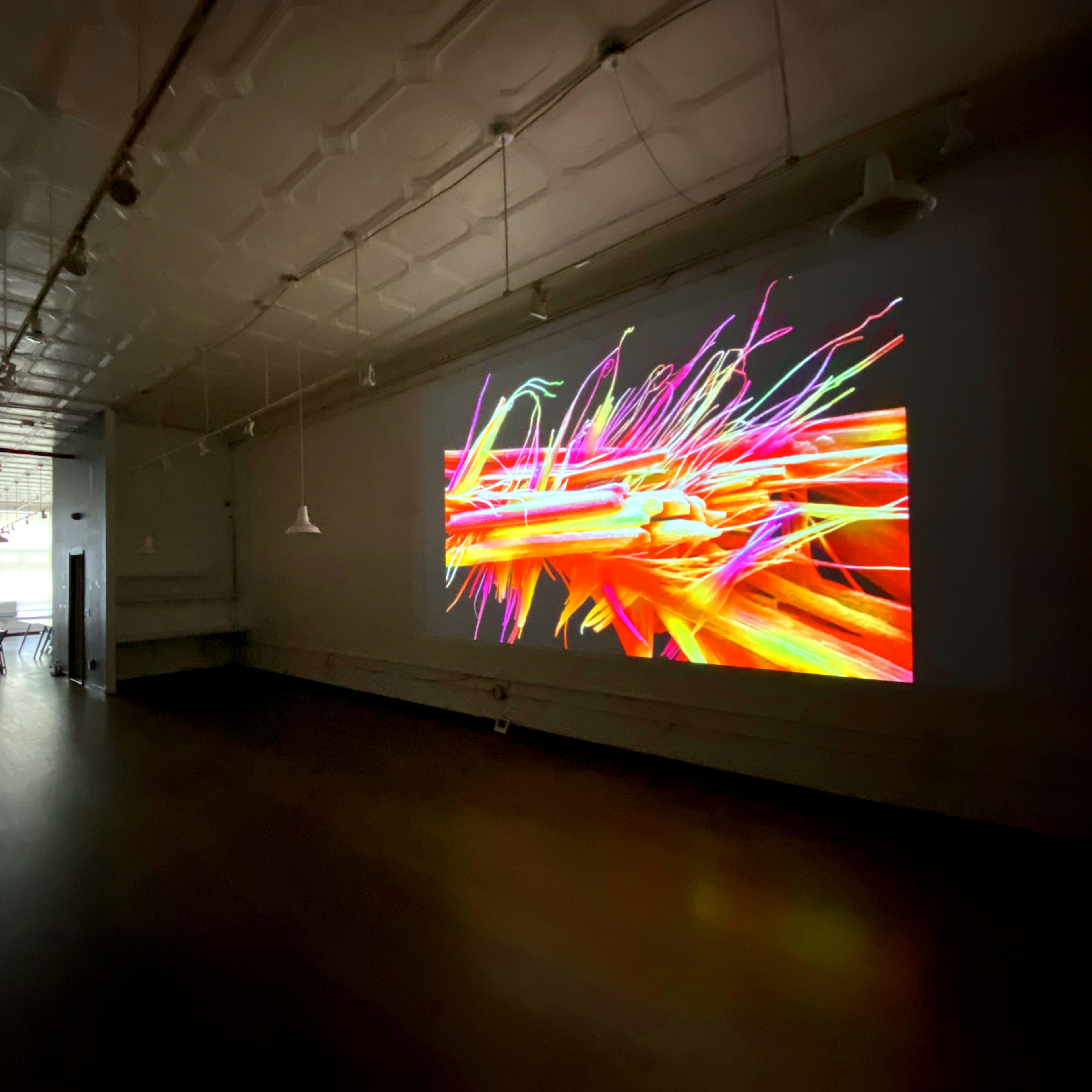 A brightly colored digital art piece is displayed on a wall in a spacious, dimly lit gallery.
