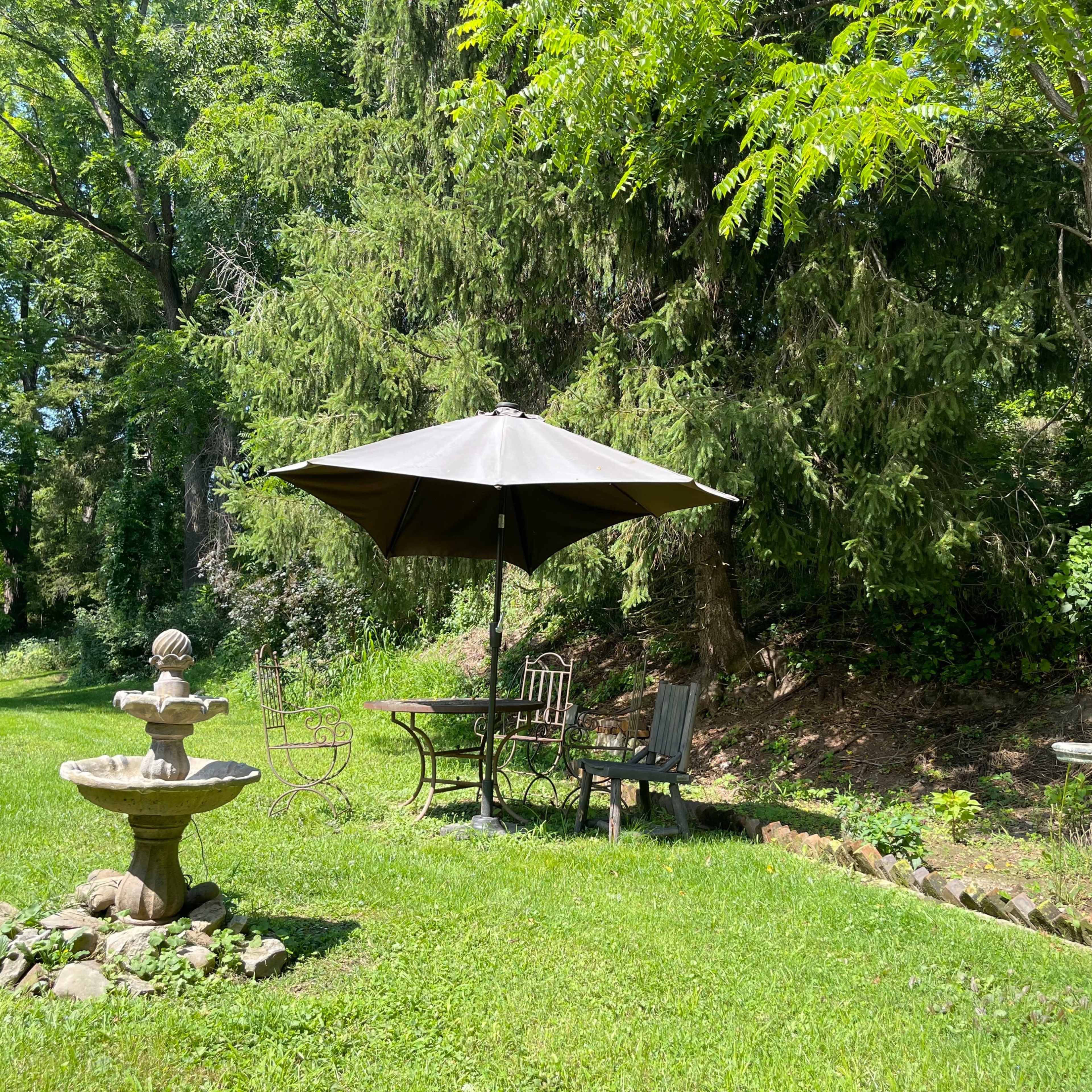 A gray patio umbrella shades a small table and chairs next to a stone fountain in a green garden.