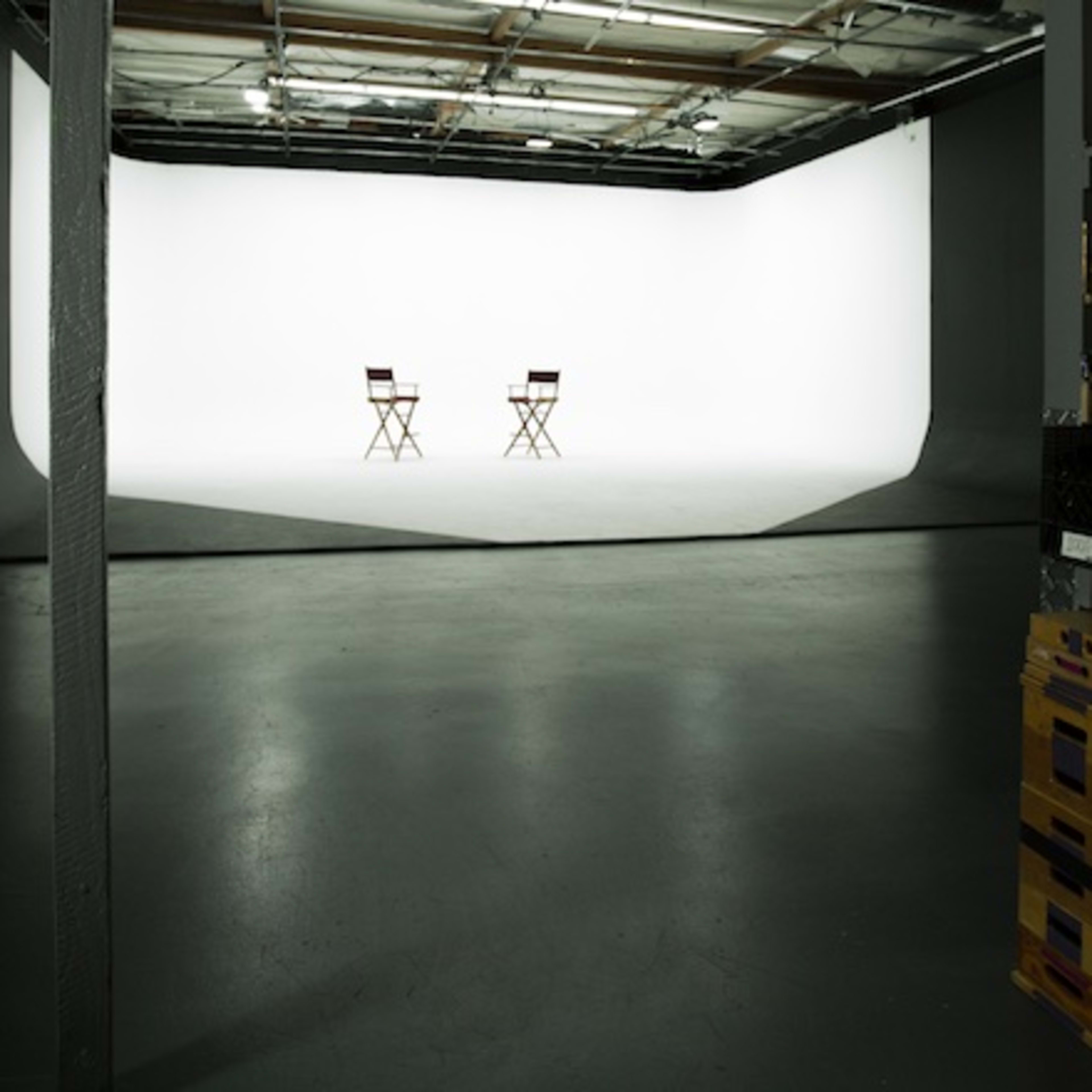 An empty studio space with two director's chairs facing a white backdrop, flanked by shelves containing equipment.