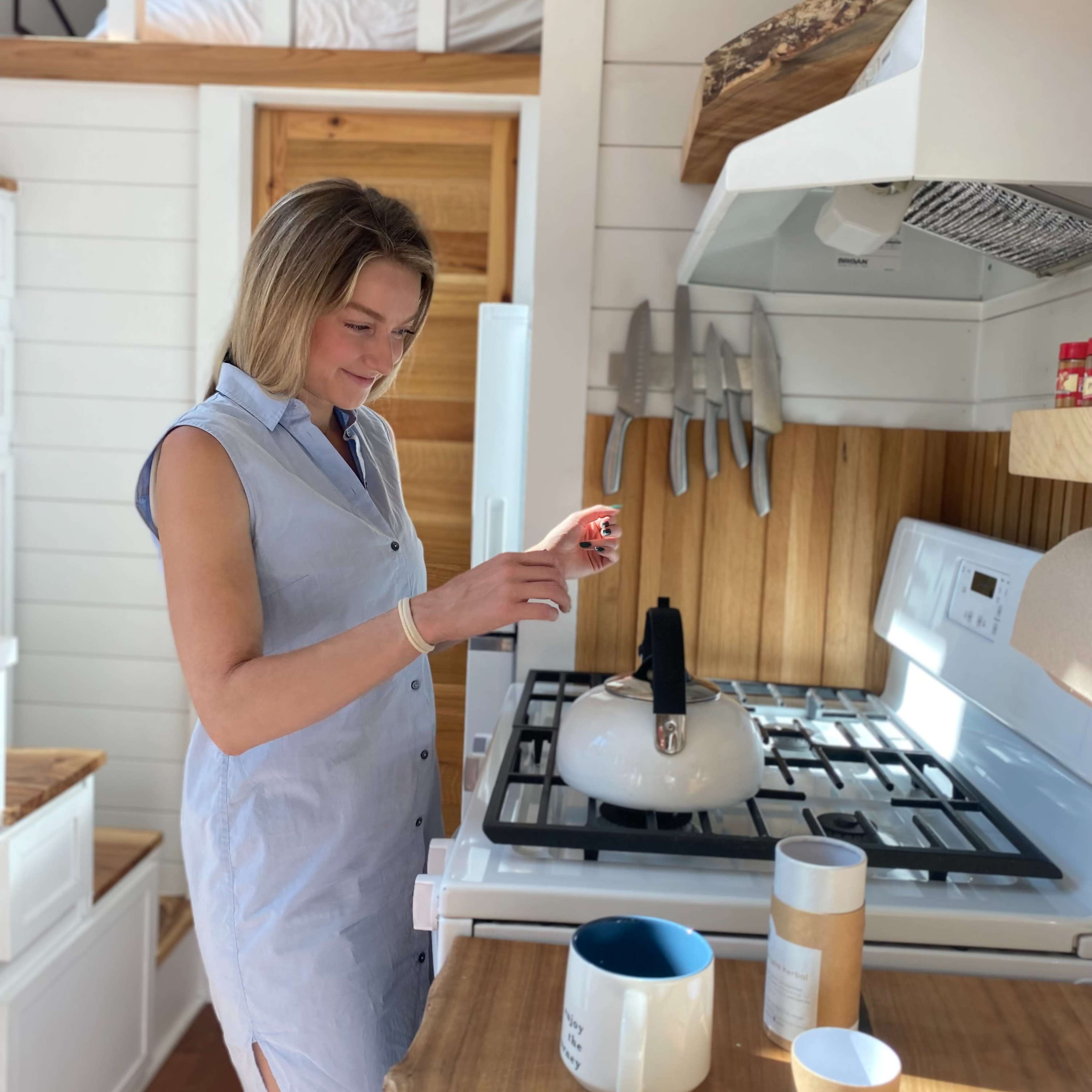 A woman stands in a bright kitchen, preparing to boil water in a kettle on the stove.
