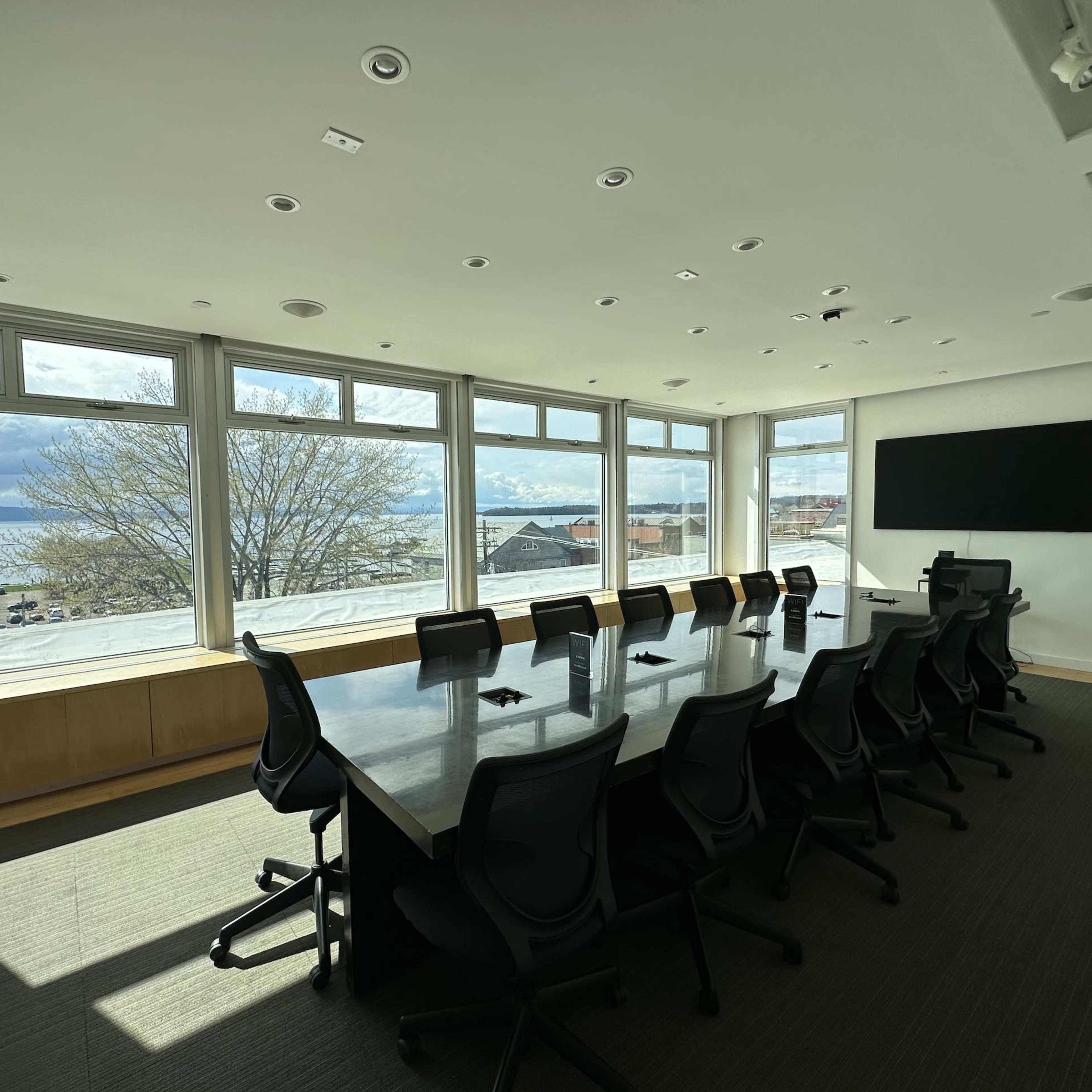 A modern conference room features a long table surrounded by chairs, with large windows offering views of the nearby waterfront and landscape.