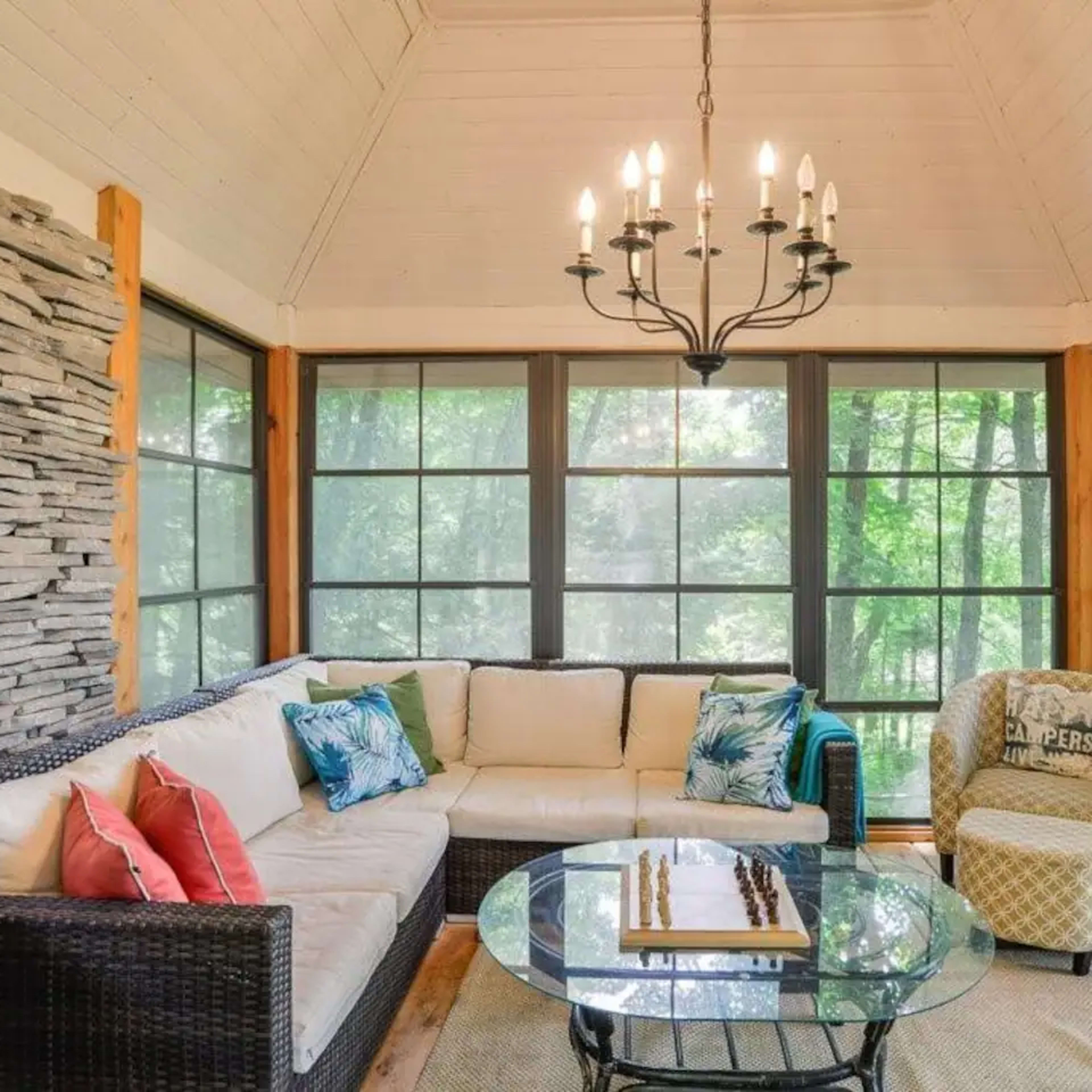 A sunroom features a sectional sofa, a glass coffee table with a chess set, and a chandelier, surrounded by large windows providing views of the outdoors.