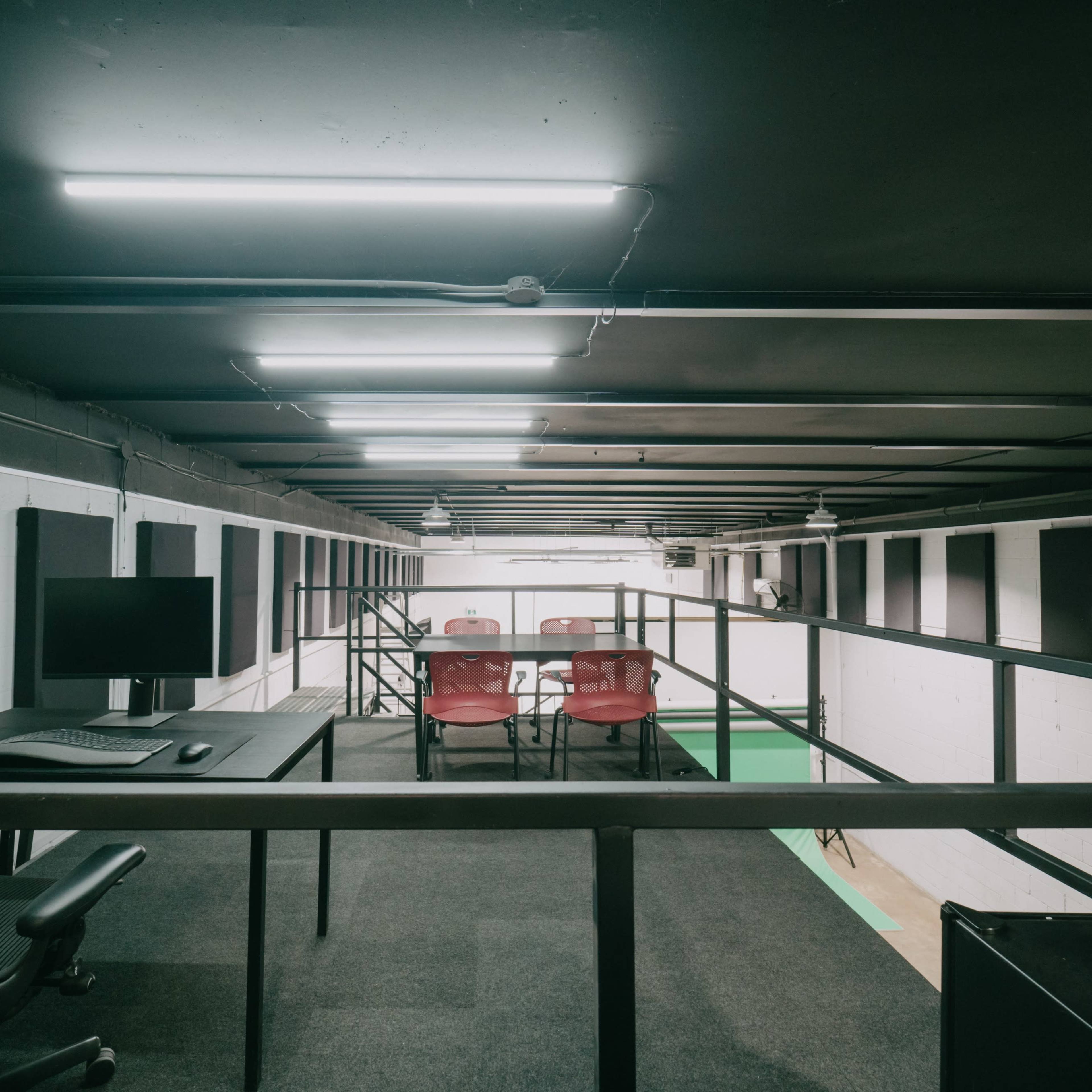 A modern office space features a raised area with a desk, computer, and red chairs arranged around a table, illuminated by overhead lighting.