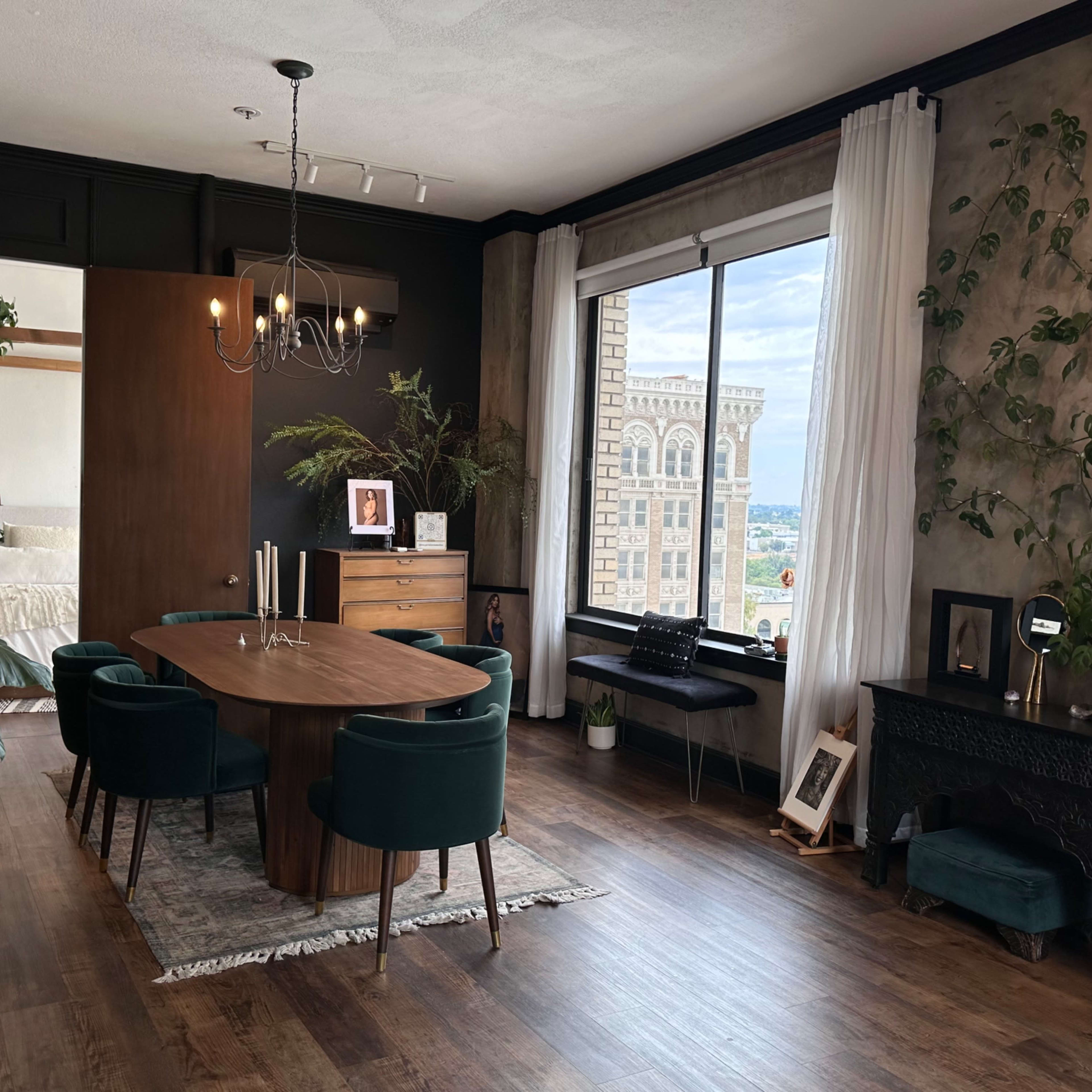 A modern dining area with a round table, green upholstered chairs, a wood dresser, and a large window overlooking a cityscape.