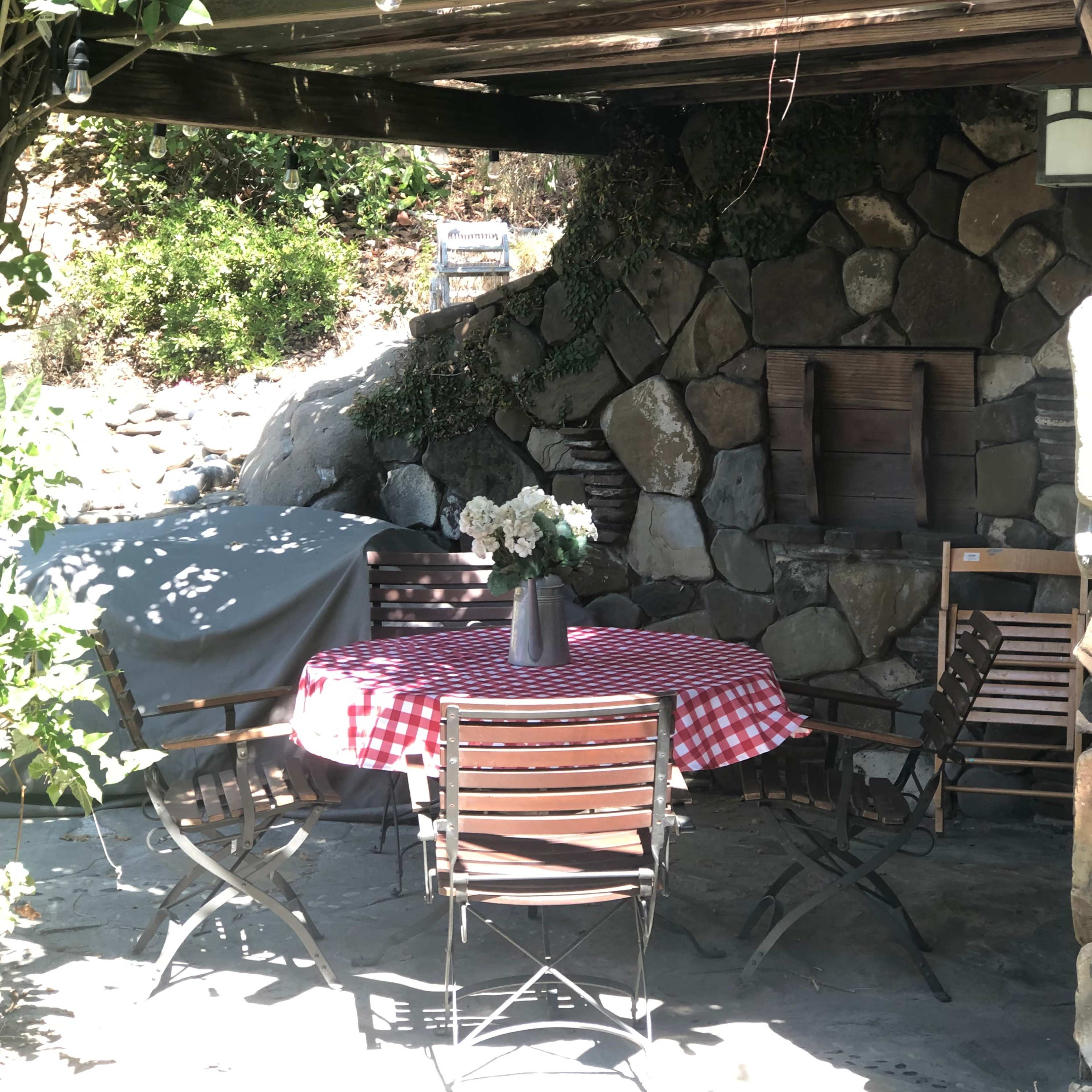 A round table with a red and white checkered tablecloth is set with a vase of flowers under a wooden patio cover surrounded by stone walls.