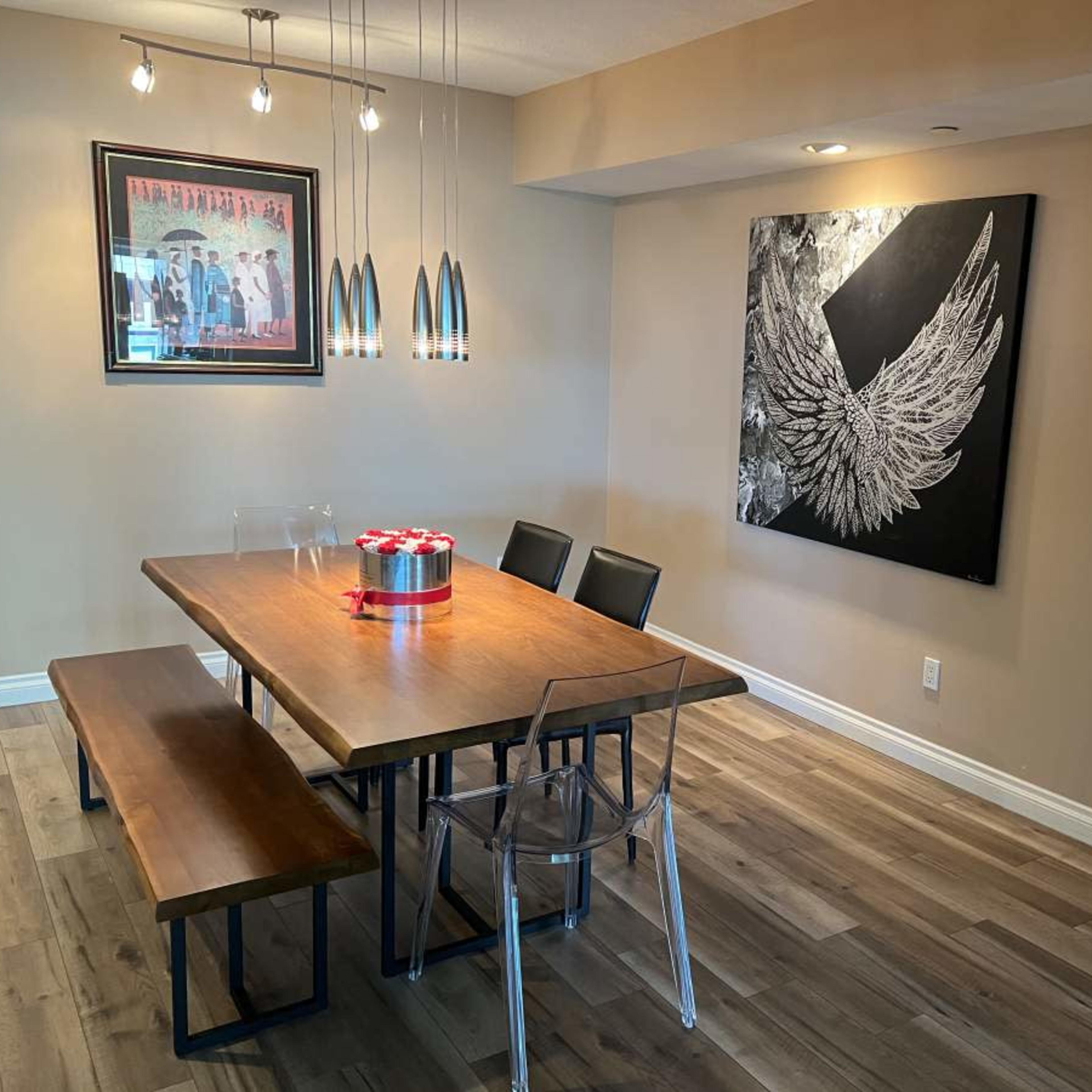 A dining area with a wooden table, clear acrylic chairs, a bench, and a large winged artwork on the wall.