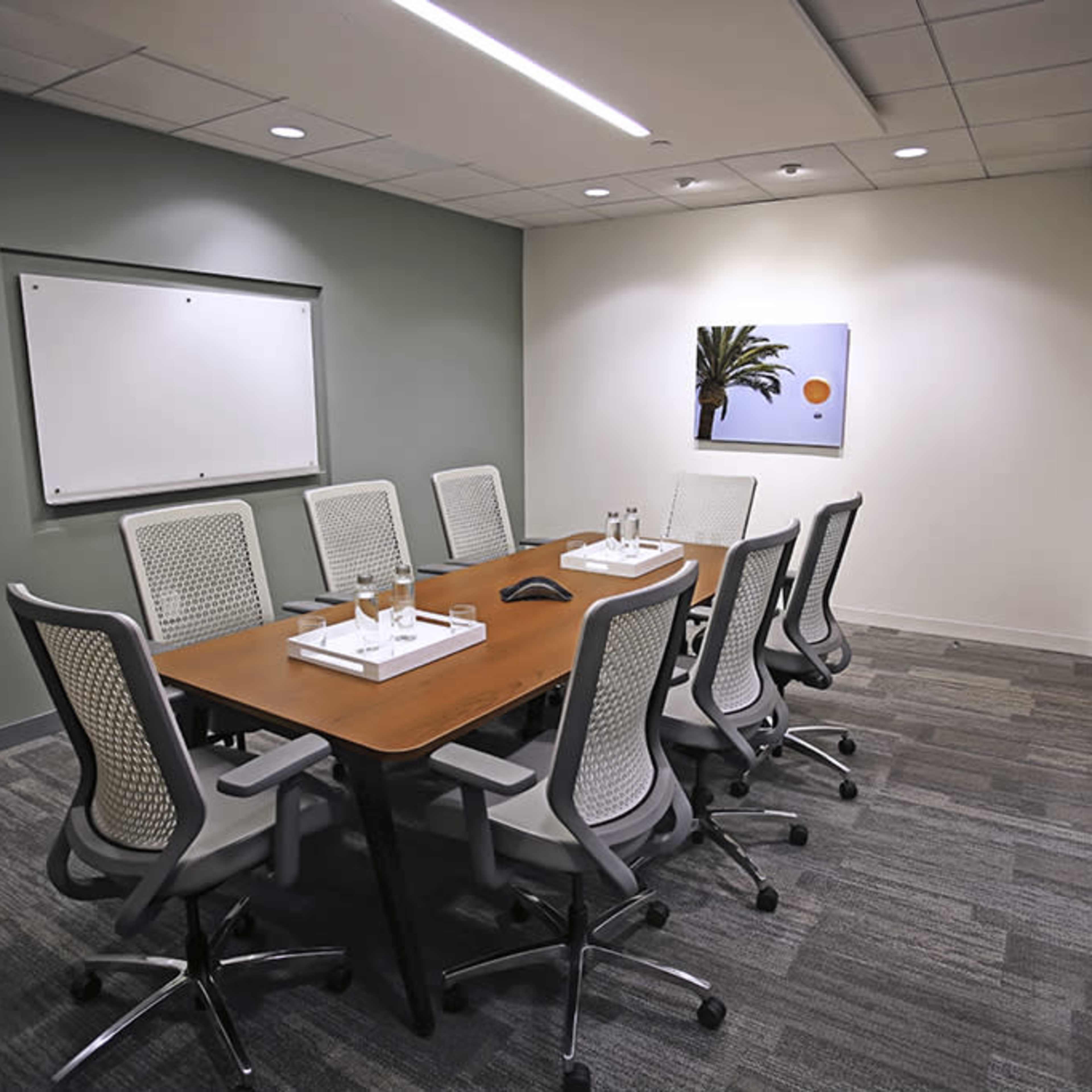 A conference room features a long wooden table surrounded by ten ergonomic chairs, with a large whiteboard and an abstract painting on the wall.