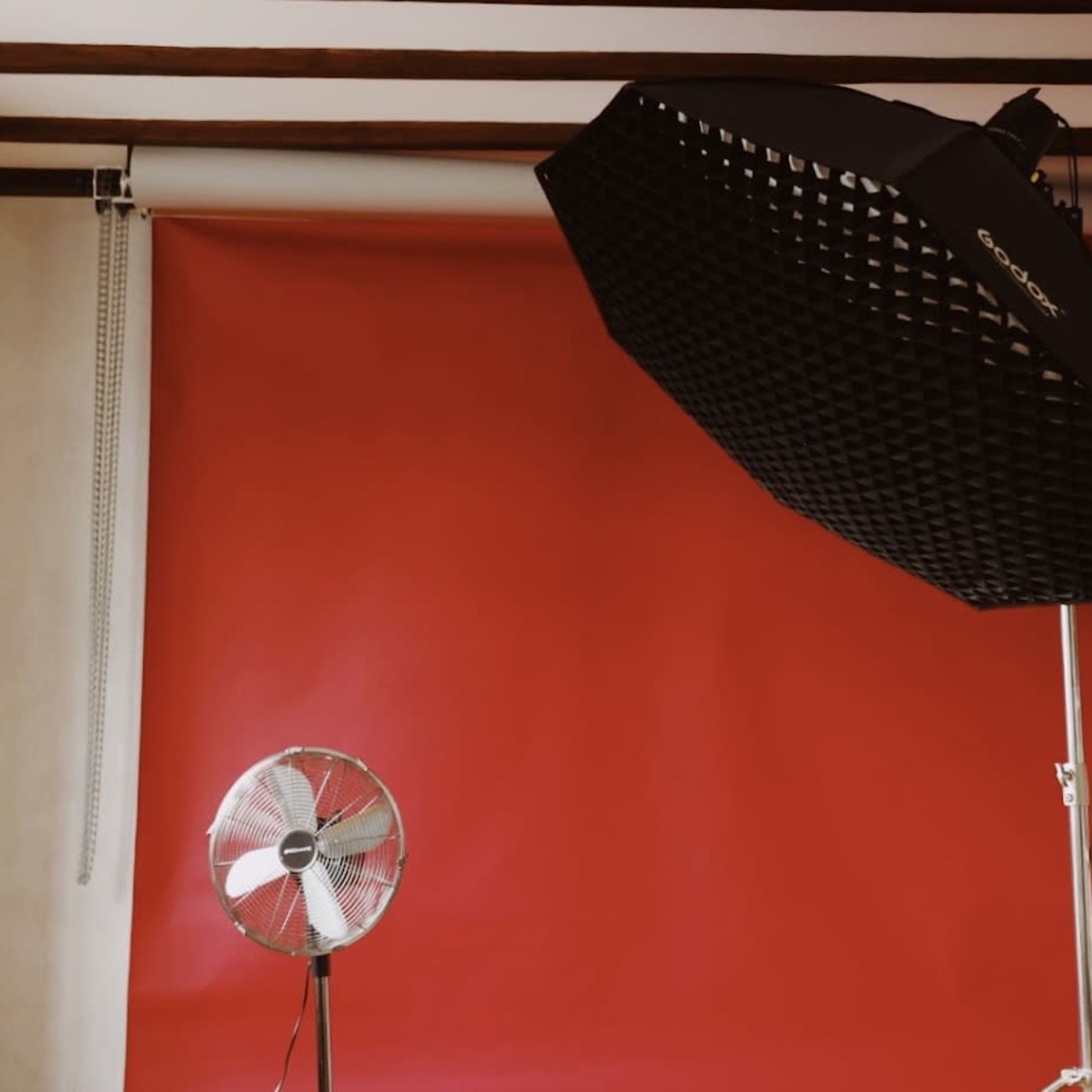 A photography studio is set up with a red backdrop, a large softbox light, and a standing fan.