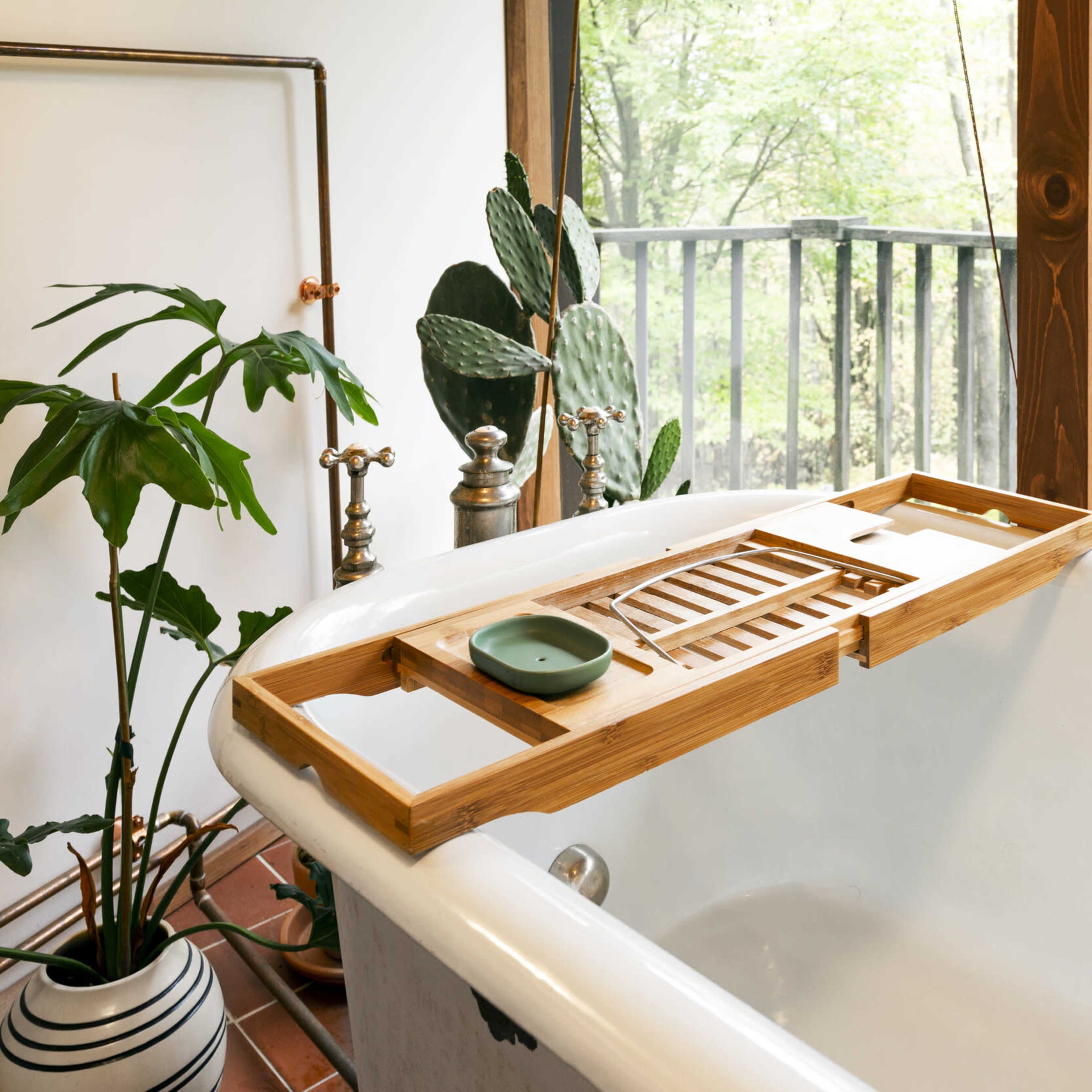 A wooden bathtub caddy rests on the edge of a white bathtub, surrounded by greenery and large windows showing a forest view.