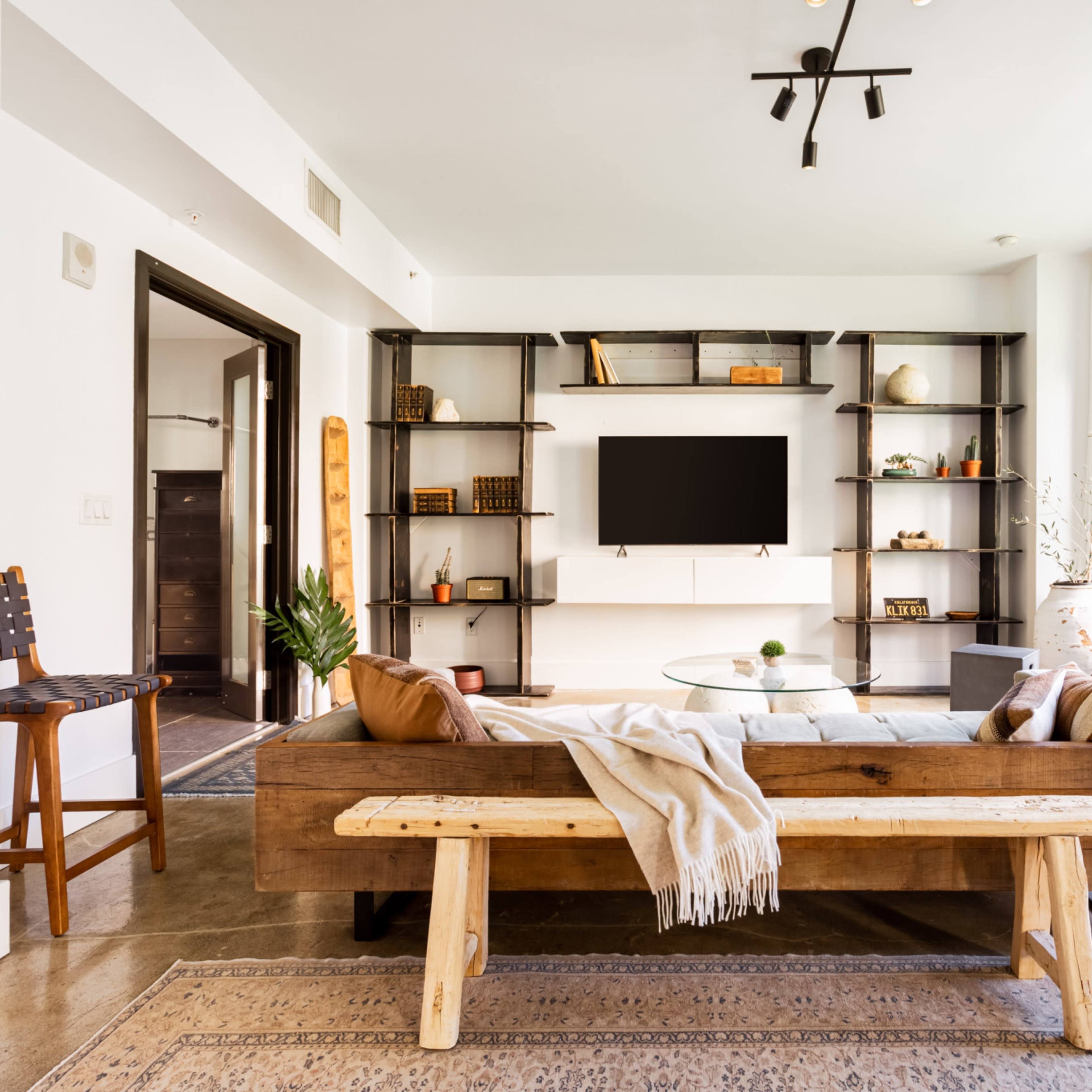 A modern living room with a wooden sofa and coffee table, a wall-mounted television, and bookshelves filled with decorative items.