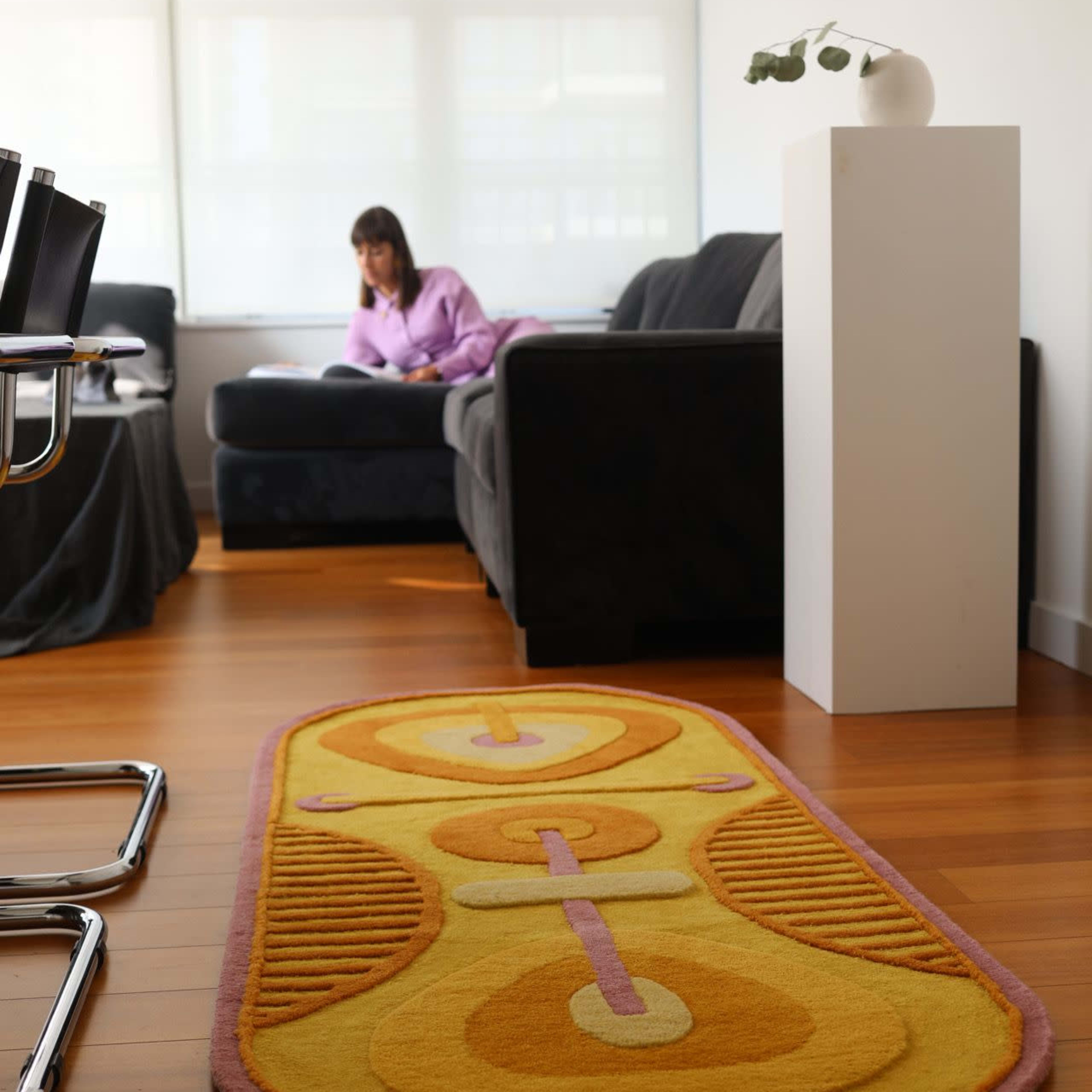 A woman sits on a dark sofa in a well-lit room with hardwood flooring, while a colorful rug runs along the floor in front of her.