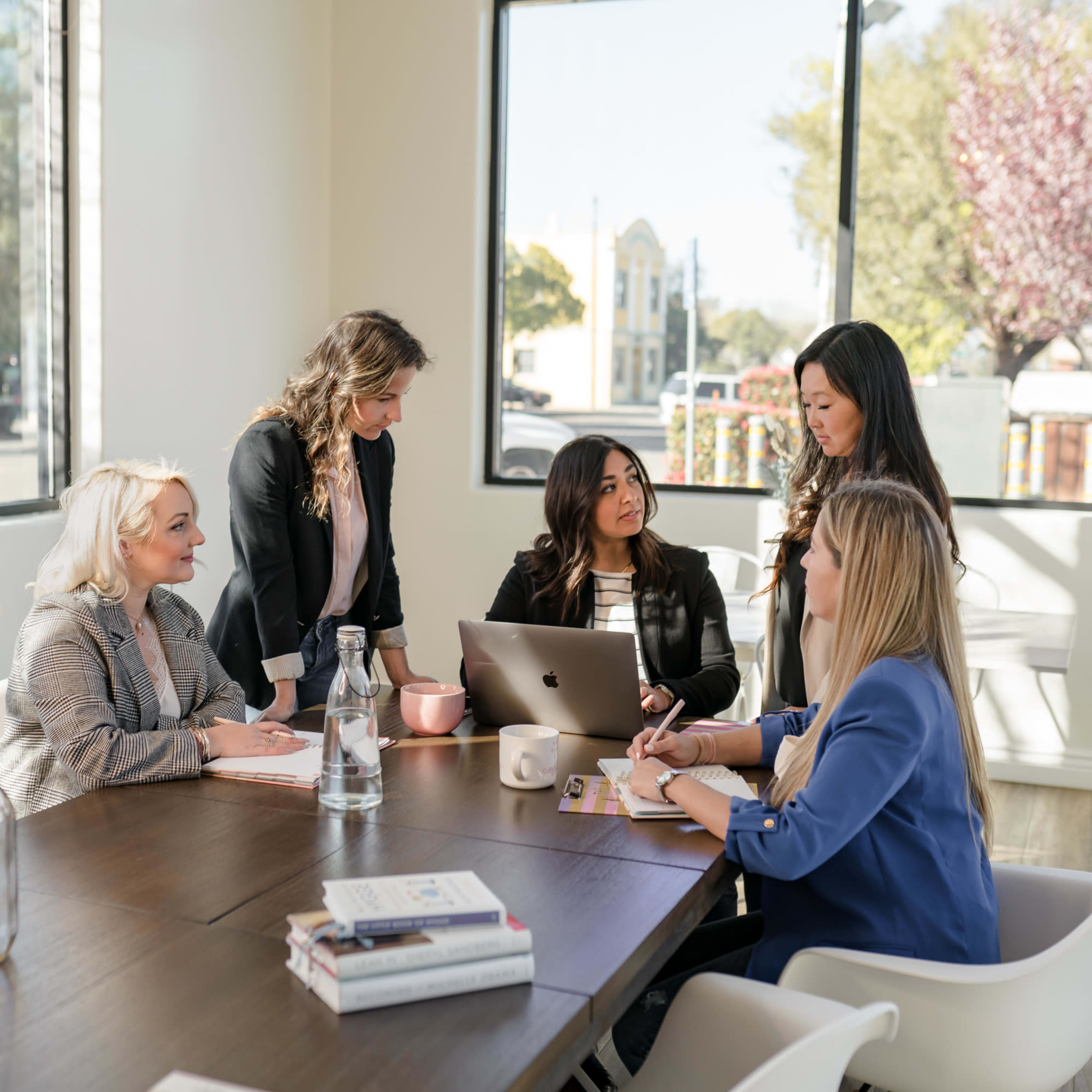 A group of six women are seated around a conference table, engaged in discussion, with laptops and notebooks visible in a bright, modern office space.