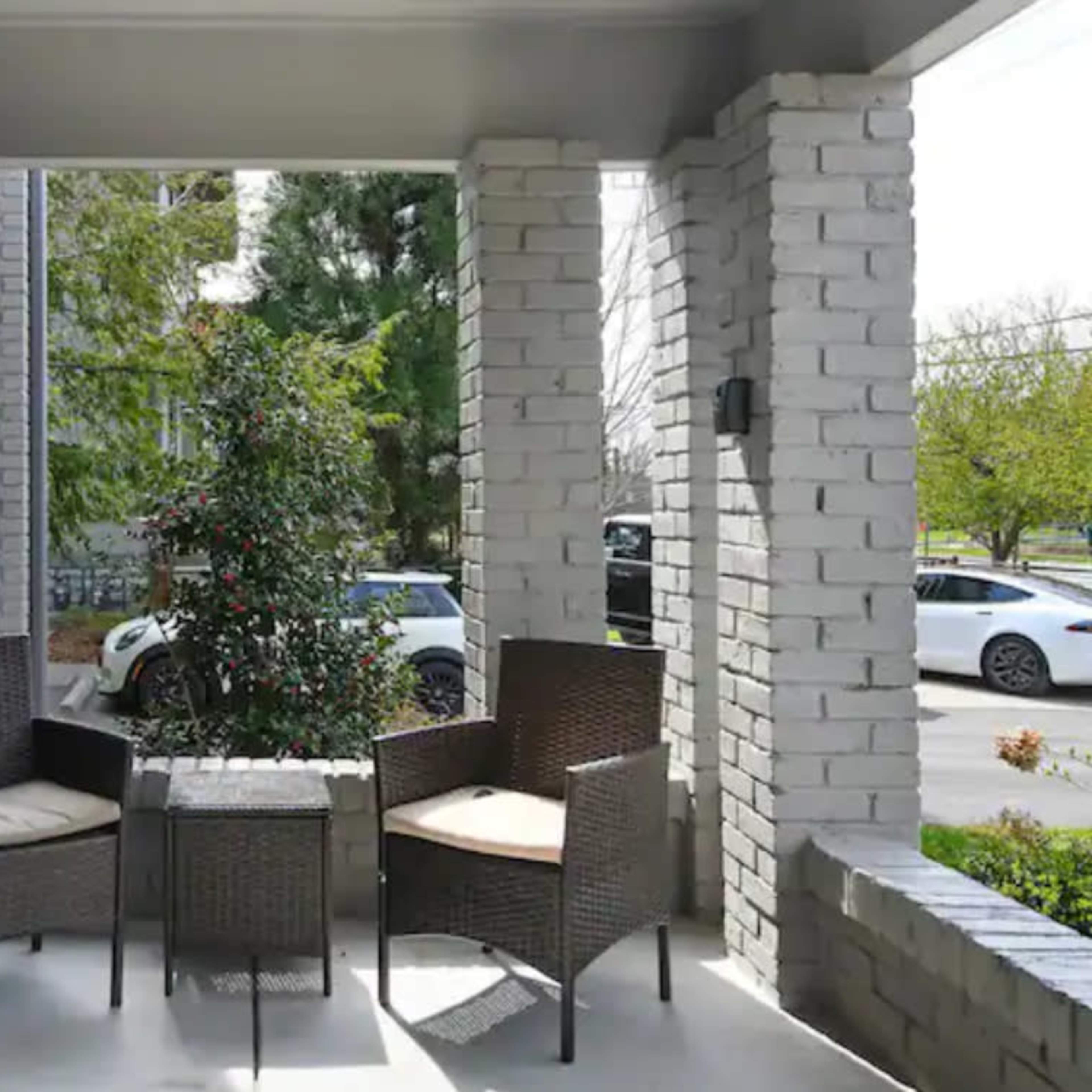 The scene shows a porch with two wicker chairs and a small table, surrounded by greenery and visible cars in the background.
