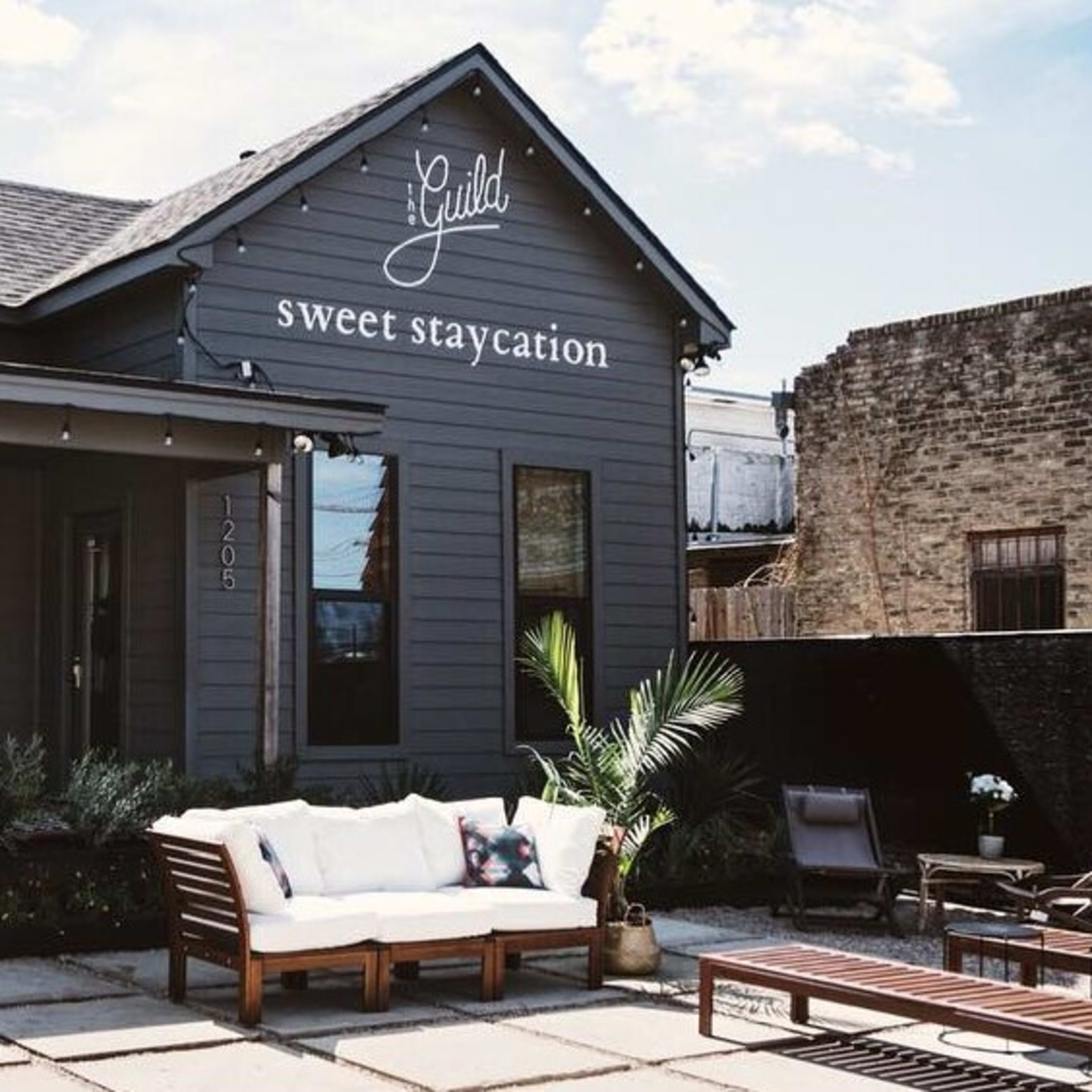 A gray house with a sign reading "sweet staycation" features a front porch with wicker chairs and a stone-paved patio.