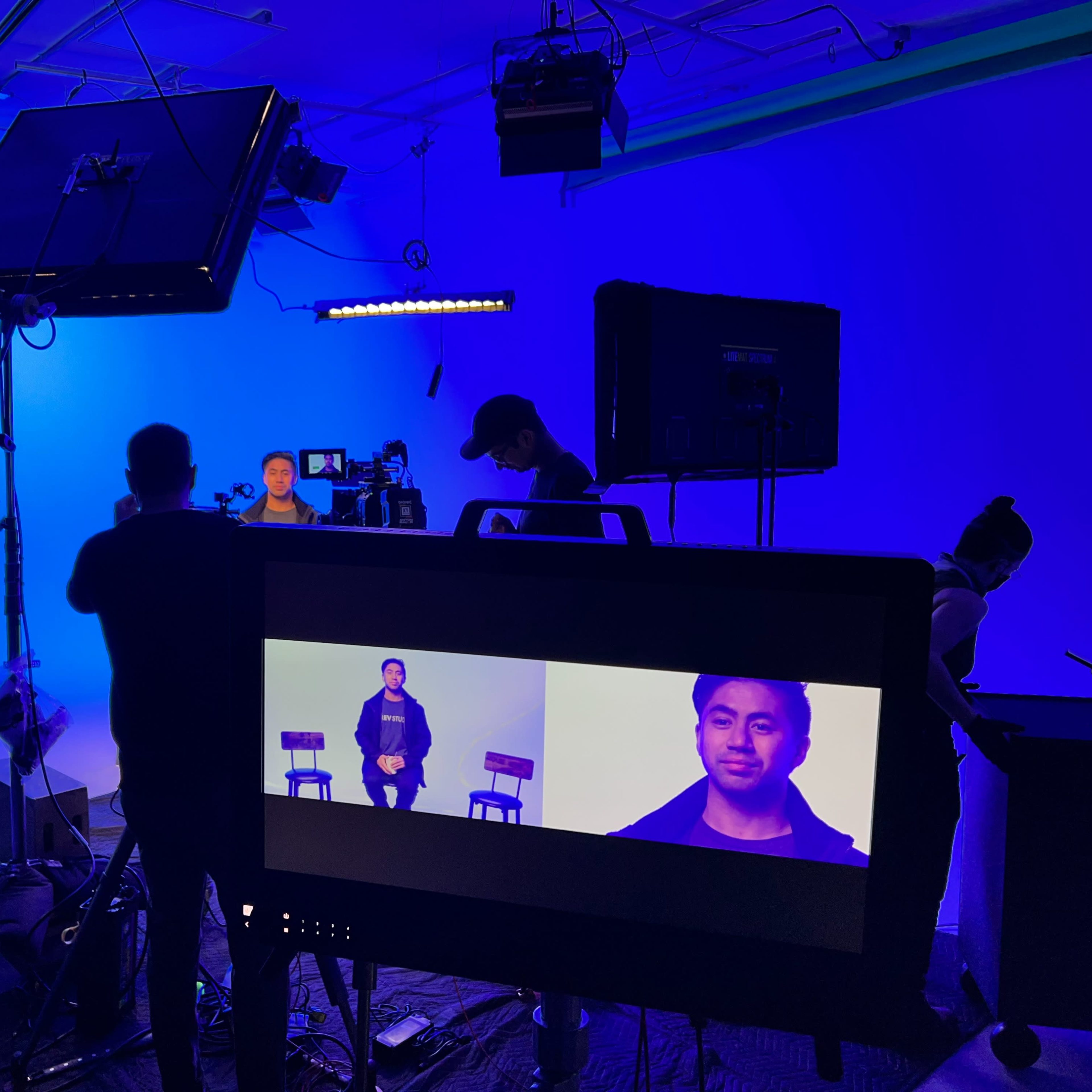 A film crew is setting up equipment in a studio with blue lighting for a video shoot featuring a man seated on a chair.
