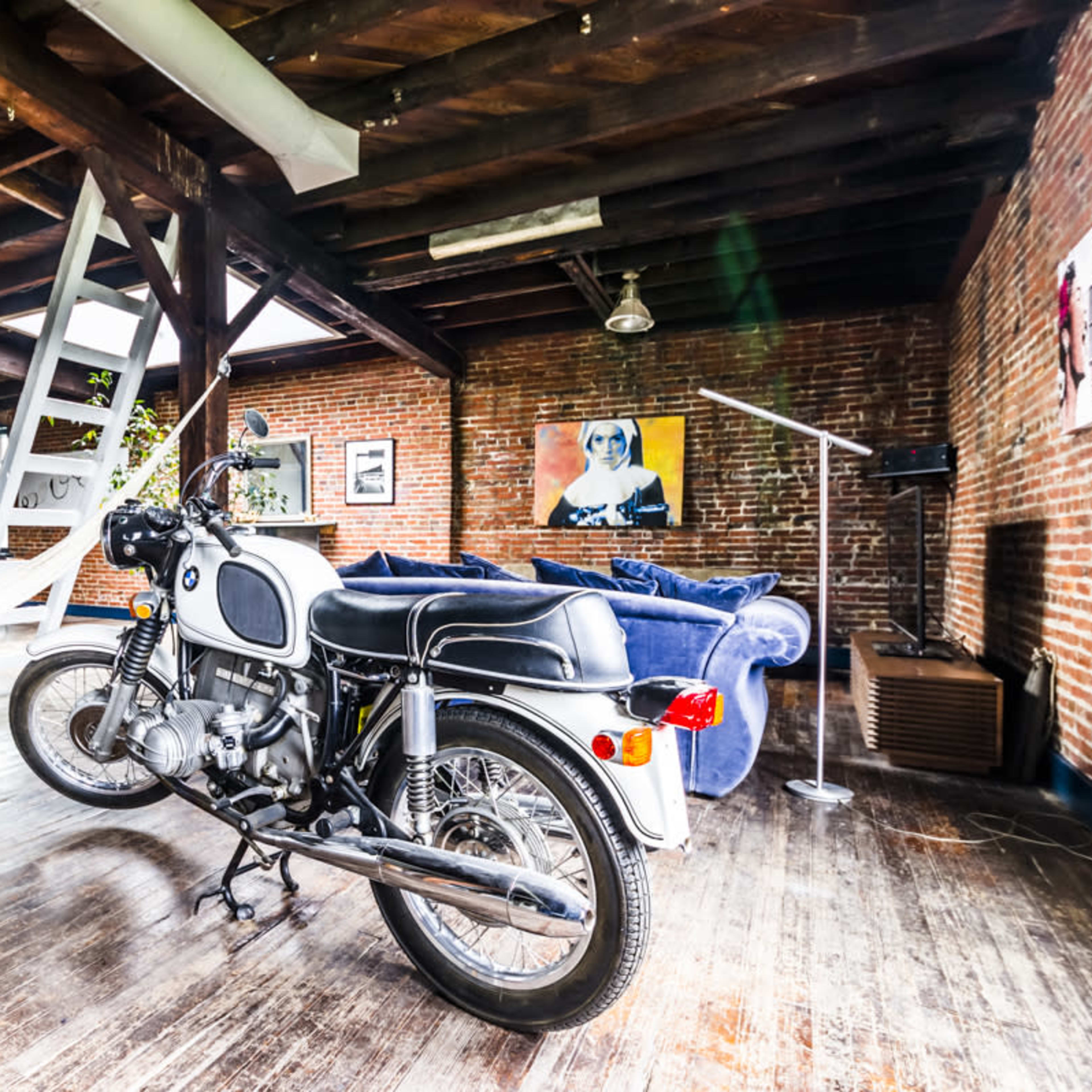 A vintage motorcycle is parked on a wooden floor in an open-concept loft with exposed brick walls and modern furnishings.