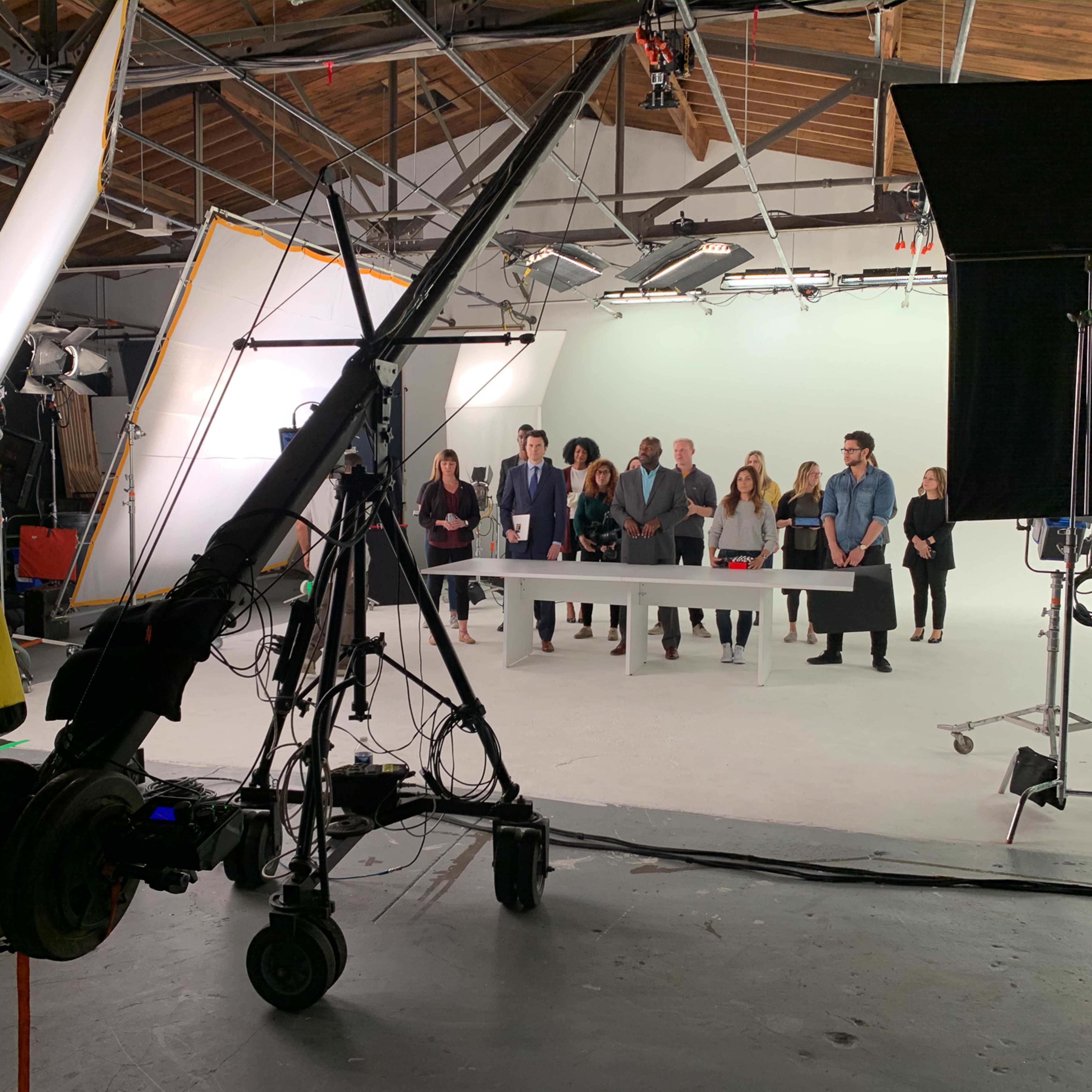 A film crew sets up equipment in a studio while a group of people stands around a table in front of a white backdrop.