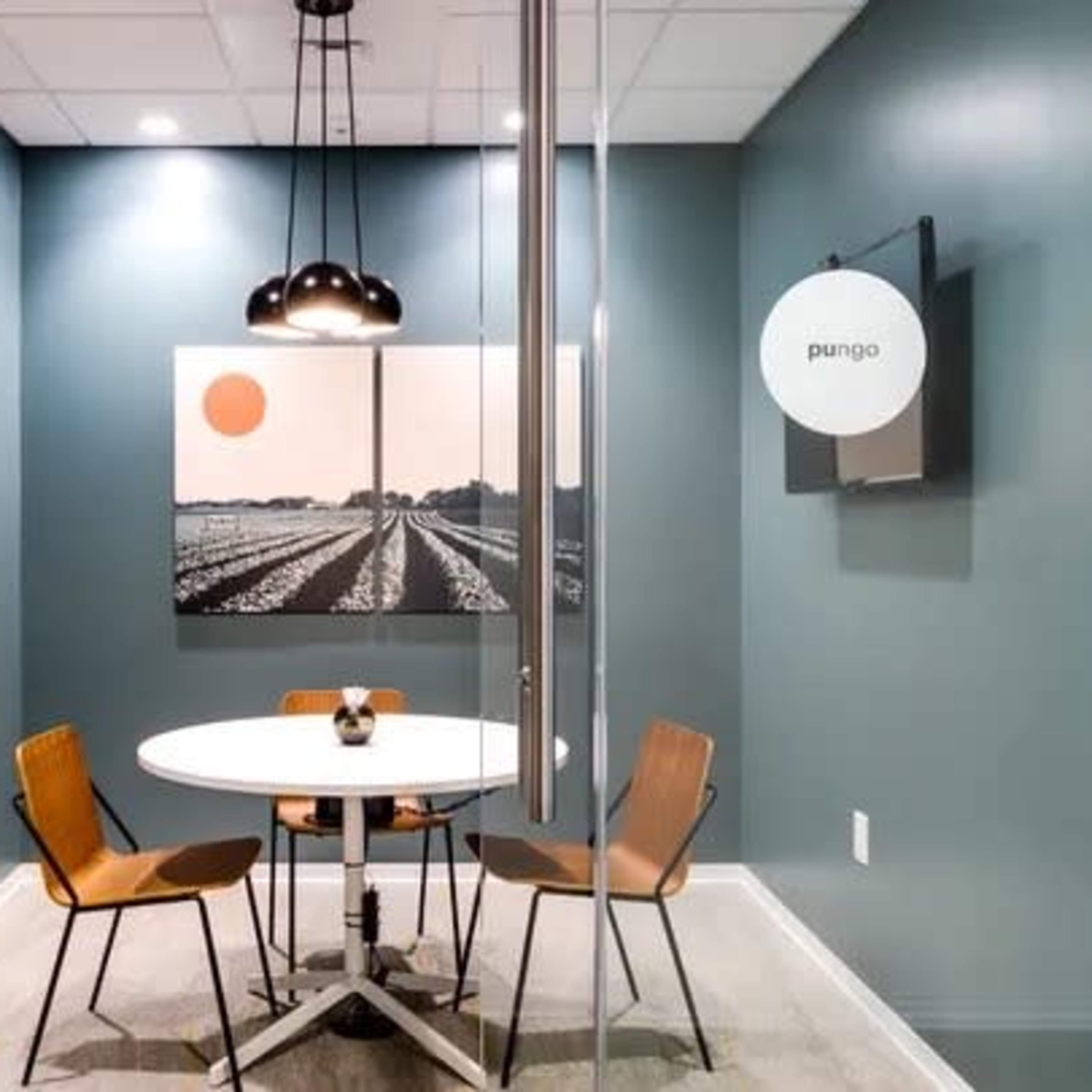 A small meeting room features a circular table with two brown chairs and a large framed artwork on the wall, illuminated by a pendant light.