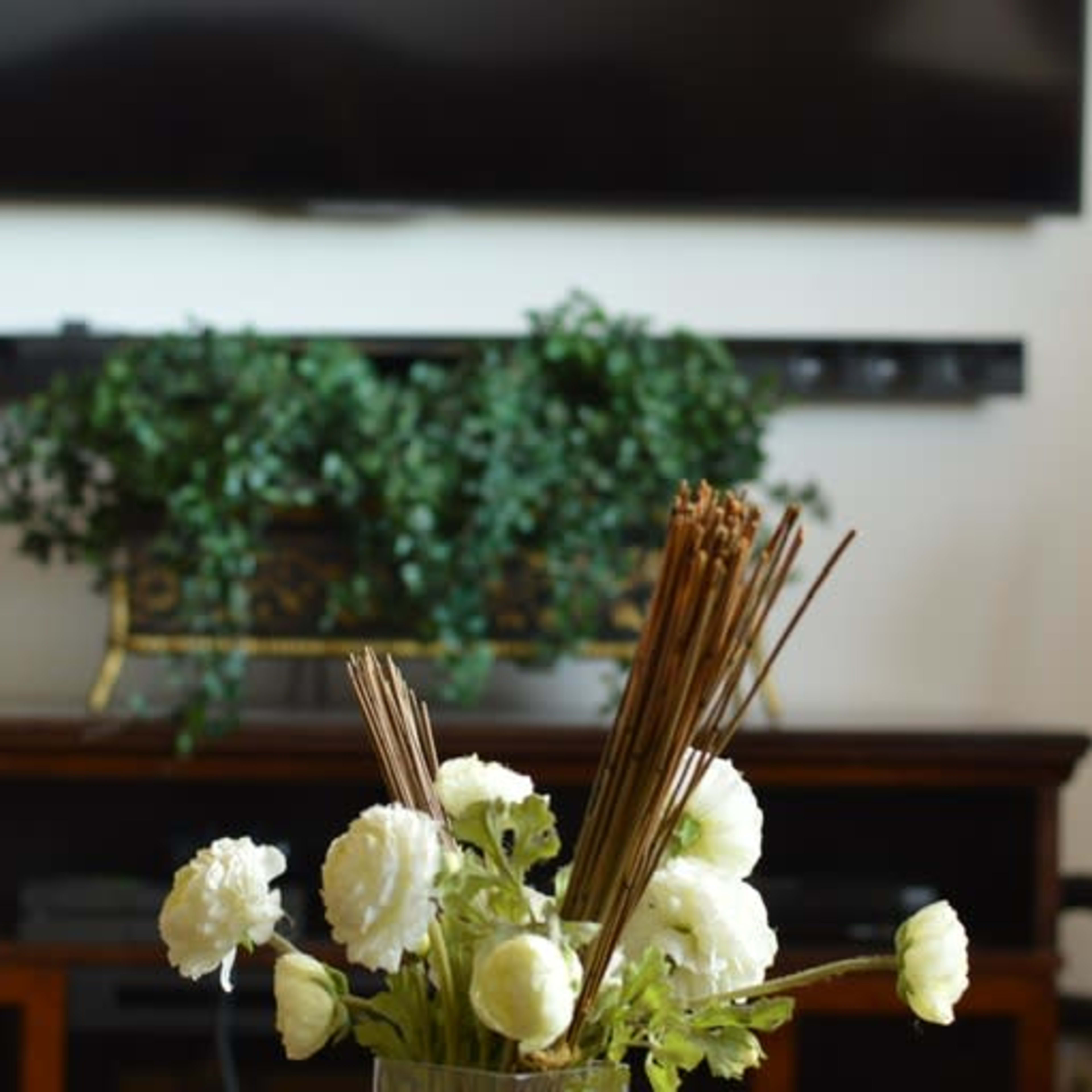 A vase with white flowers and decorative sticks sits on a table in front of a television and a stand with plants.